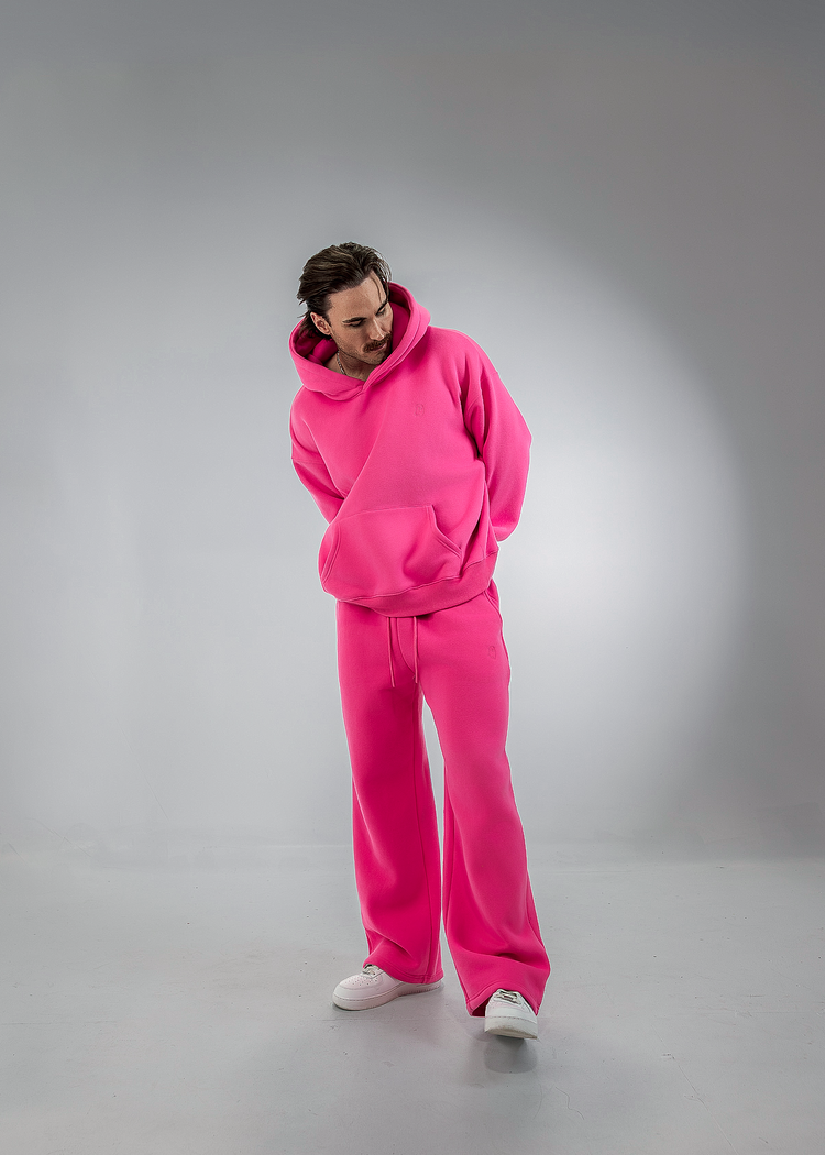 A man wearing a matching bright pink fleece tracksuit, including a pullover hoodie and wide-leg sweatpants, standing against a gray studio background.
