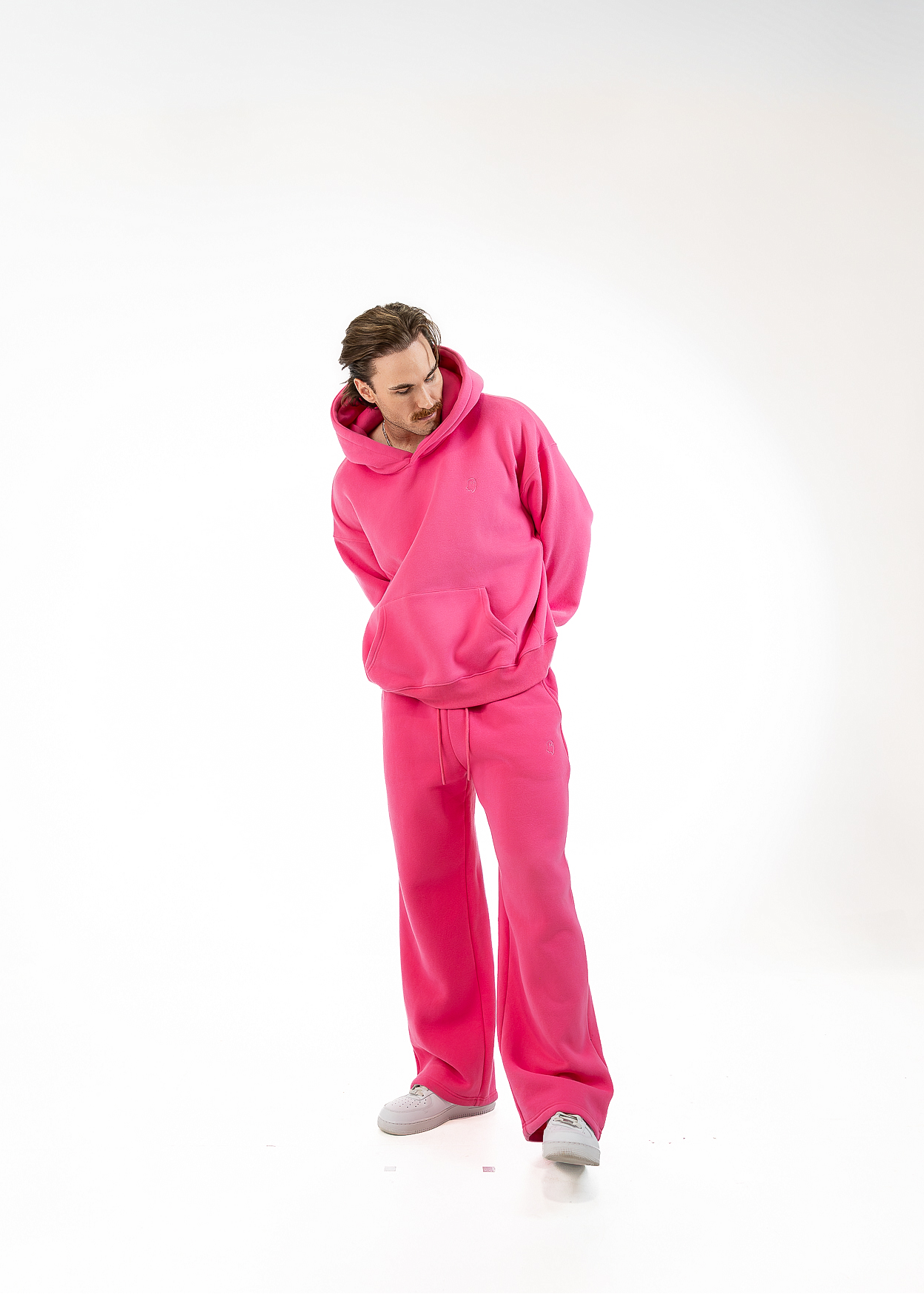 A man wearing a bright pink fleece tracksuit consisting of a pullover hoodie and matching wide-leg sweatpants, standing against a seamless white studio background.