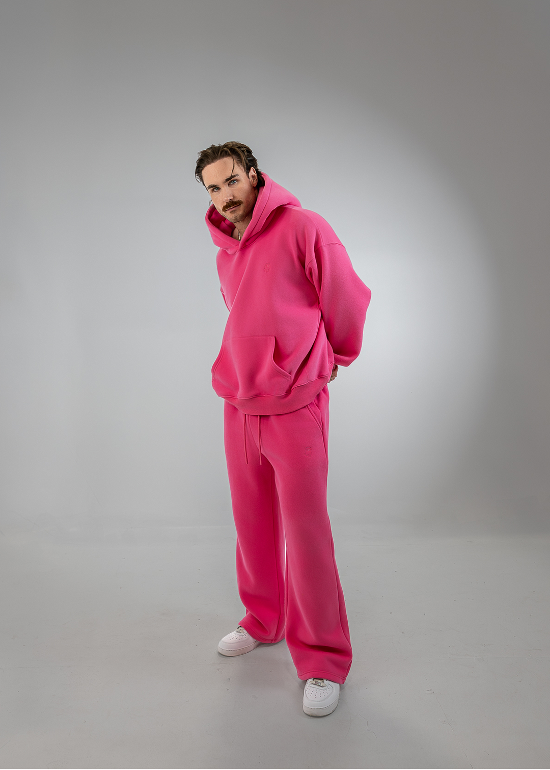 A man wearing a bright pink matching lounge set consisting of a hoodie and wide-leg sweatpants, paired with white sneakers, standing against a gray studio background.