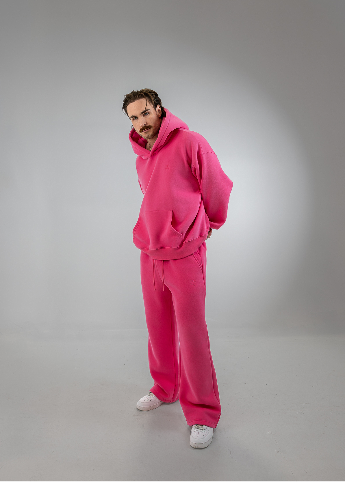 A man wearing a bright pink matching lounge set consisting of a hoodie and wide-leg sweatpants, paired with white sneakers, standing against a gray studio background.