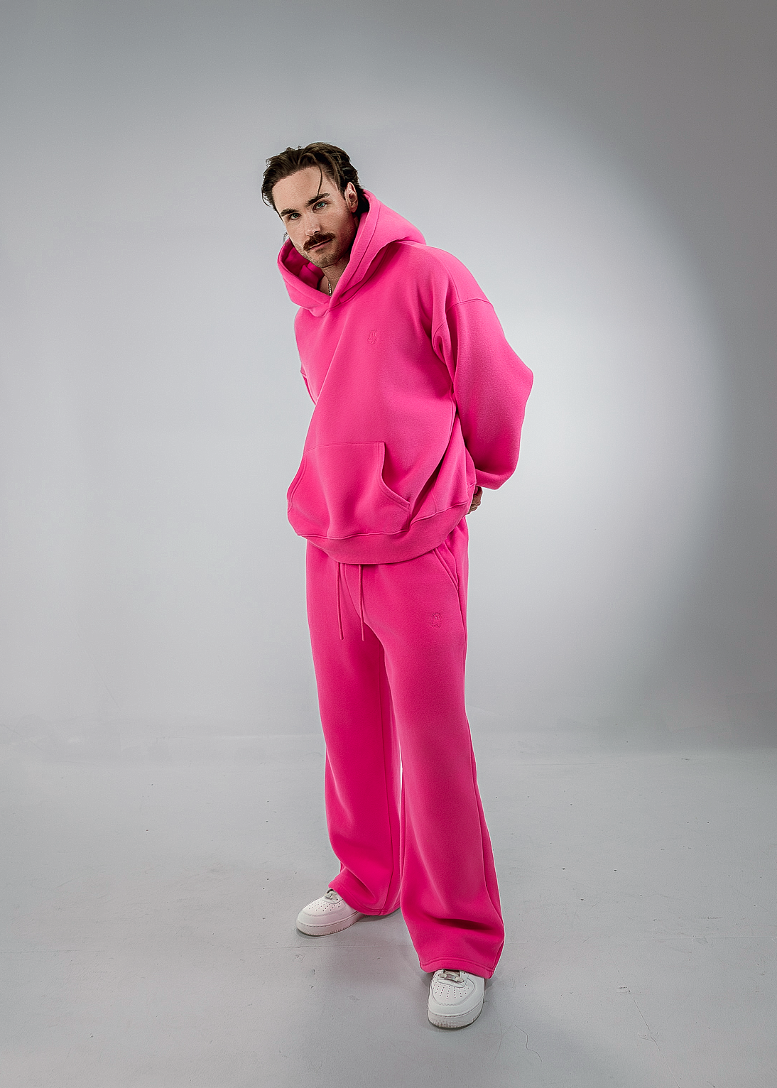 A man wearing a bright pink matching lounge set consisting of a pullover hoodie and wide-leg sweatpants, paired with white sneakers, standing in a studio setting.