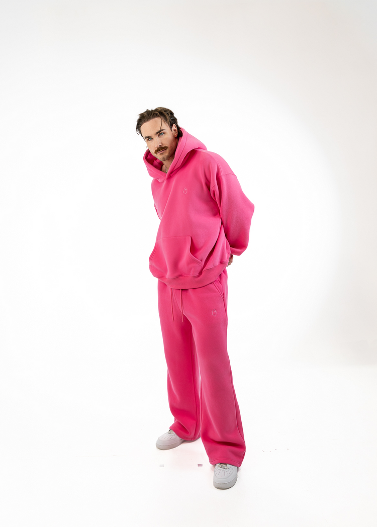 A man wearing a bright pink matching hoodie and sweatpants set, standing against a plain white studio background.
