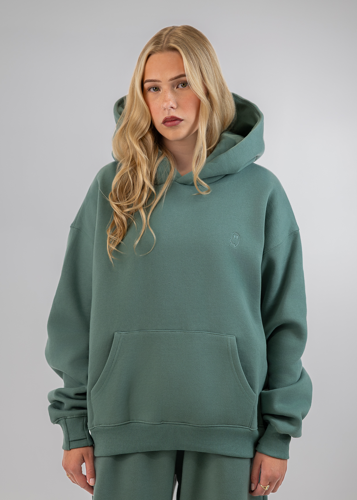 A blonde woman wearing an oversized sage green pullover hoodie with a kangaroo pocket and matching sweatpants, photographed against a neutral gray studio background.