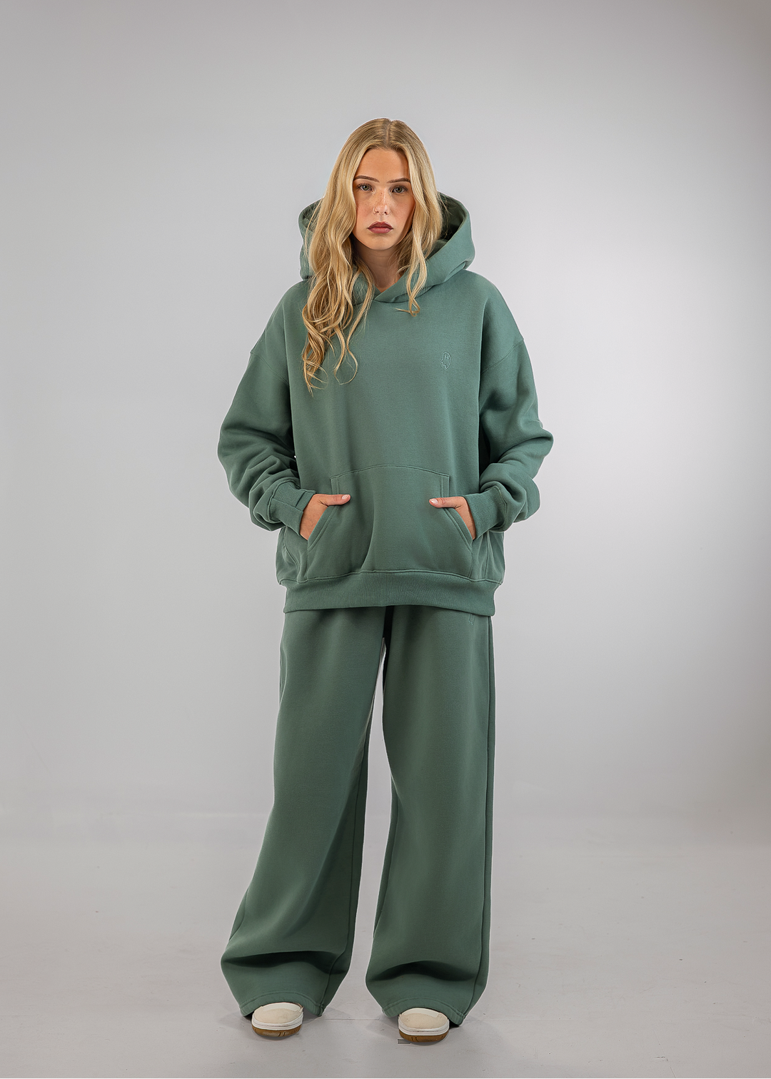 A woman modeling a matching sage green lounge set consisting of an oversized pullover hoodie and wide-leg sweatpants, standing against a neutral gray studio background.