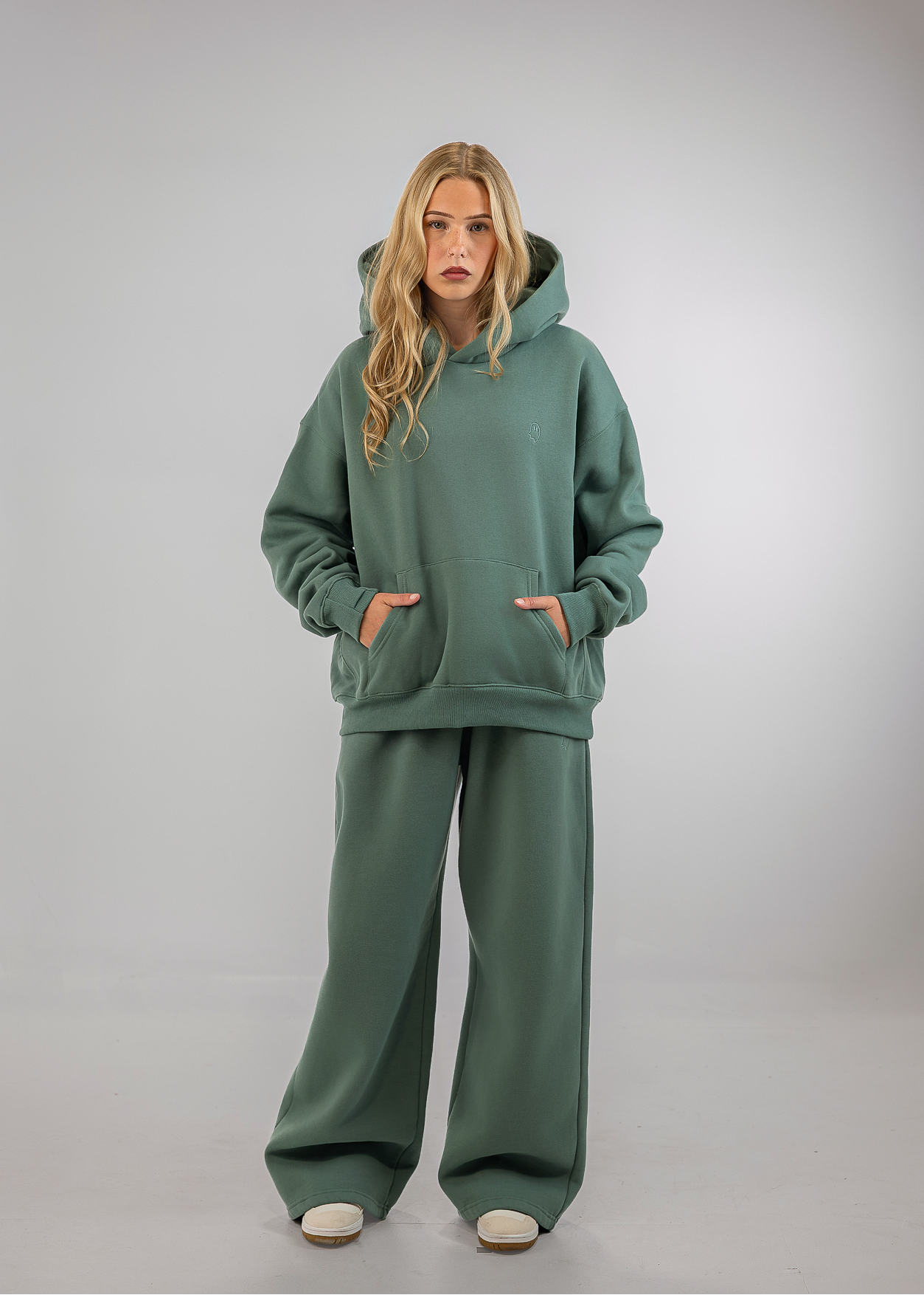 A woman modeling a matching sage green lounge set consisting of an oversized pullover hoodie and wide-leg sweatpants, standing against a neutral gray studio background.