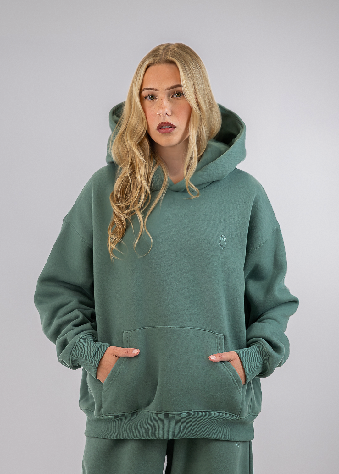 A blonde woman wearing a sage green pullover hoodie and matching sweatpants, standing against a light gray studio background.
