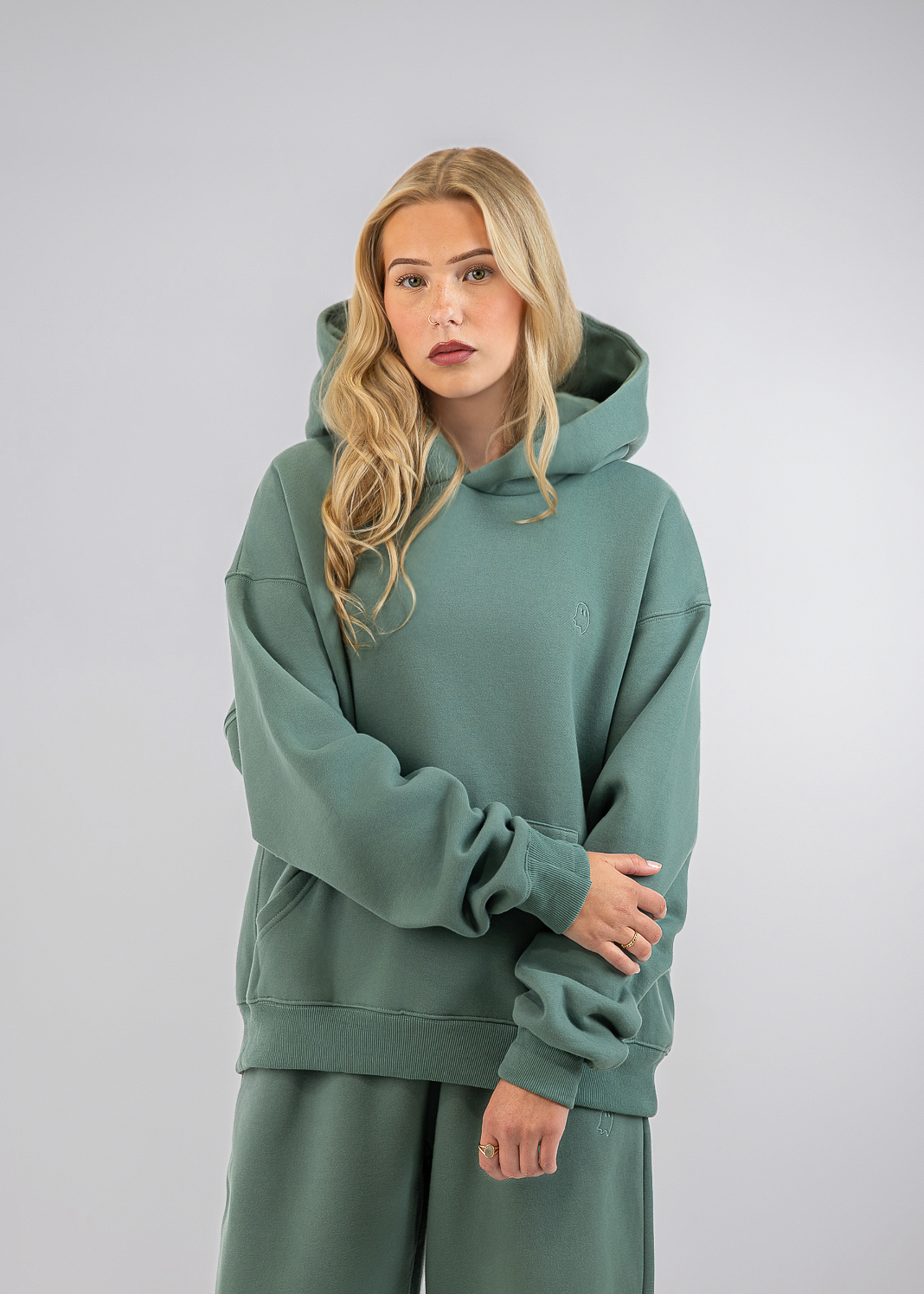 A blonde woman modeling a matching sage green hoodie and wide-leg sweatpants set against a neutral gray studio background.