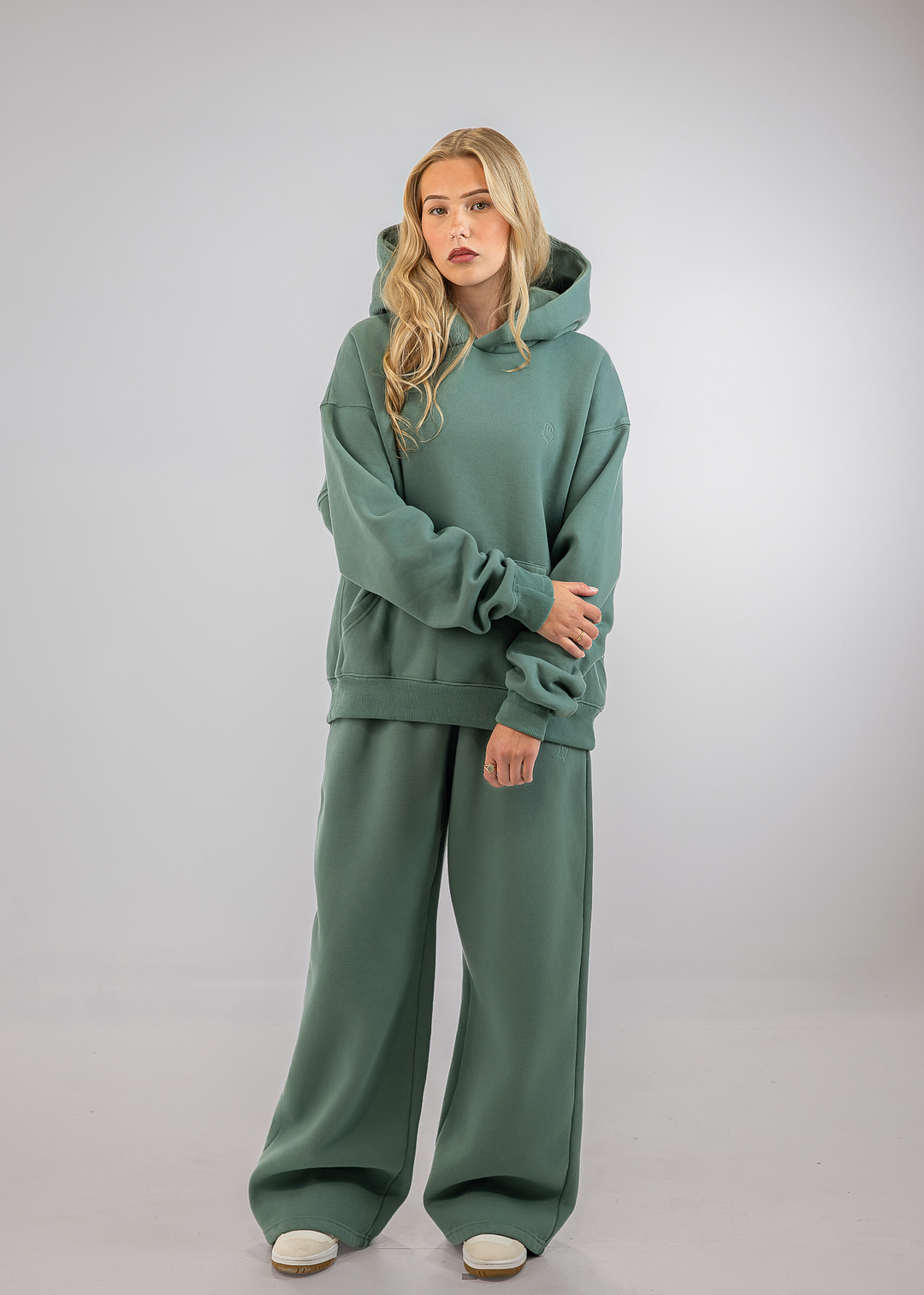 A woman modeling a matching sage green loungewear set consisting of an oversized pullover hoodie and wide-leg sweatpants, photographed against a neutral light gray studio background.