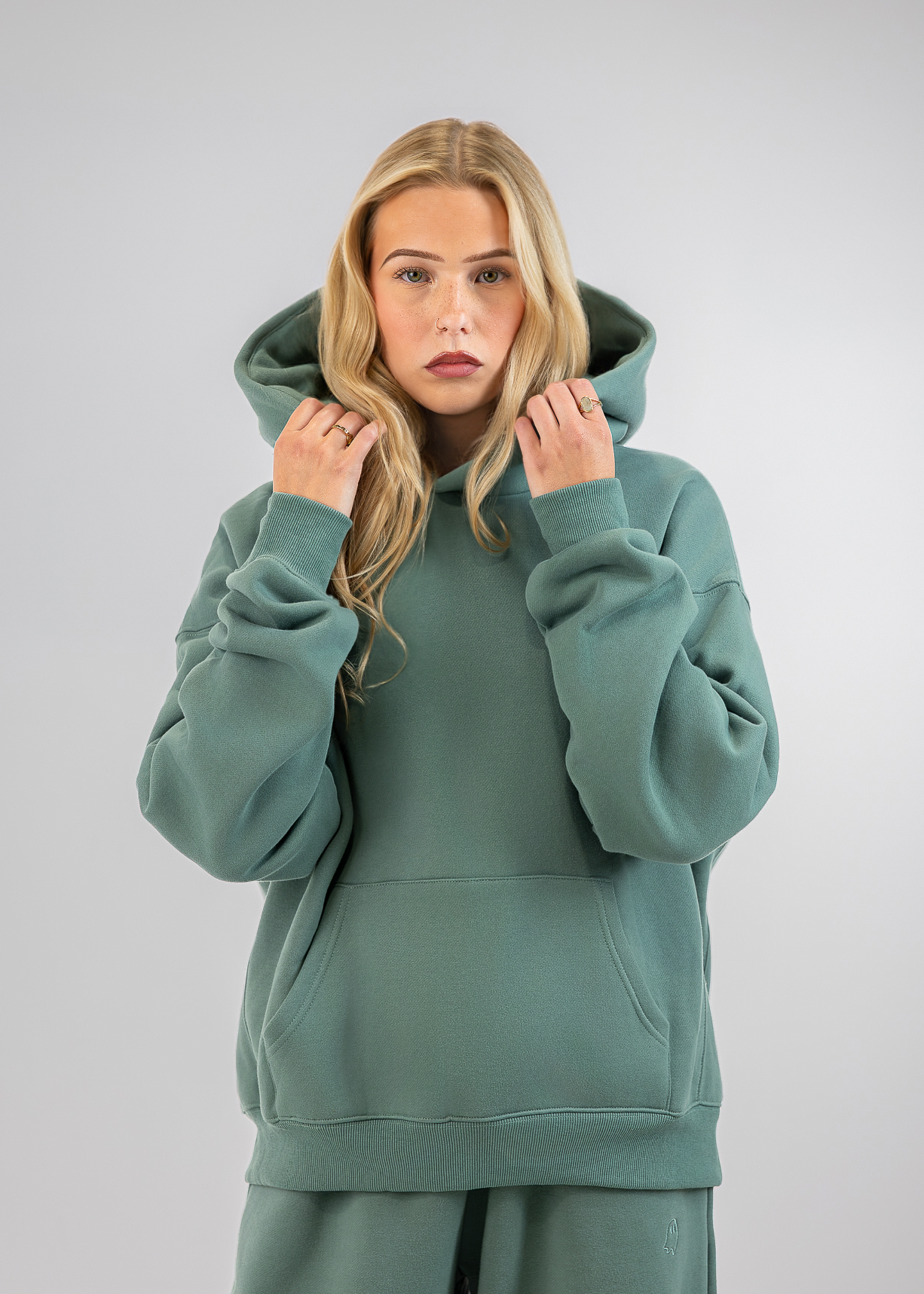 A blonde woman wearing a sage green pullover hoodie and matching sweatpants, posing against a neutral light gray studio background.