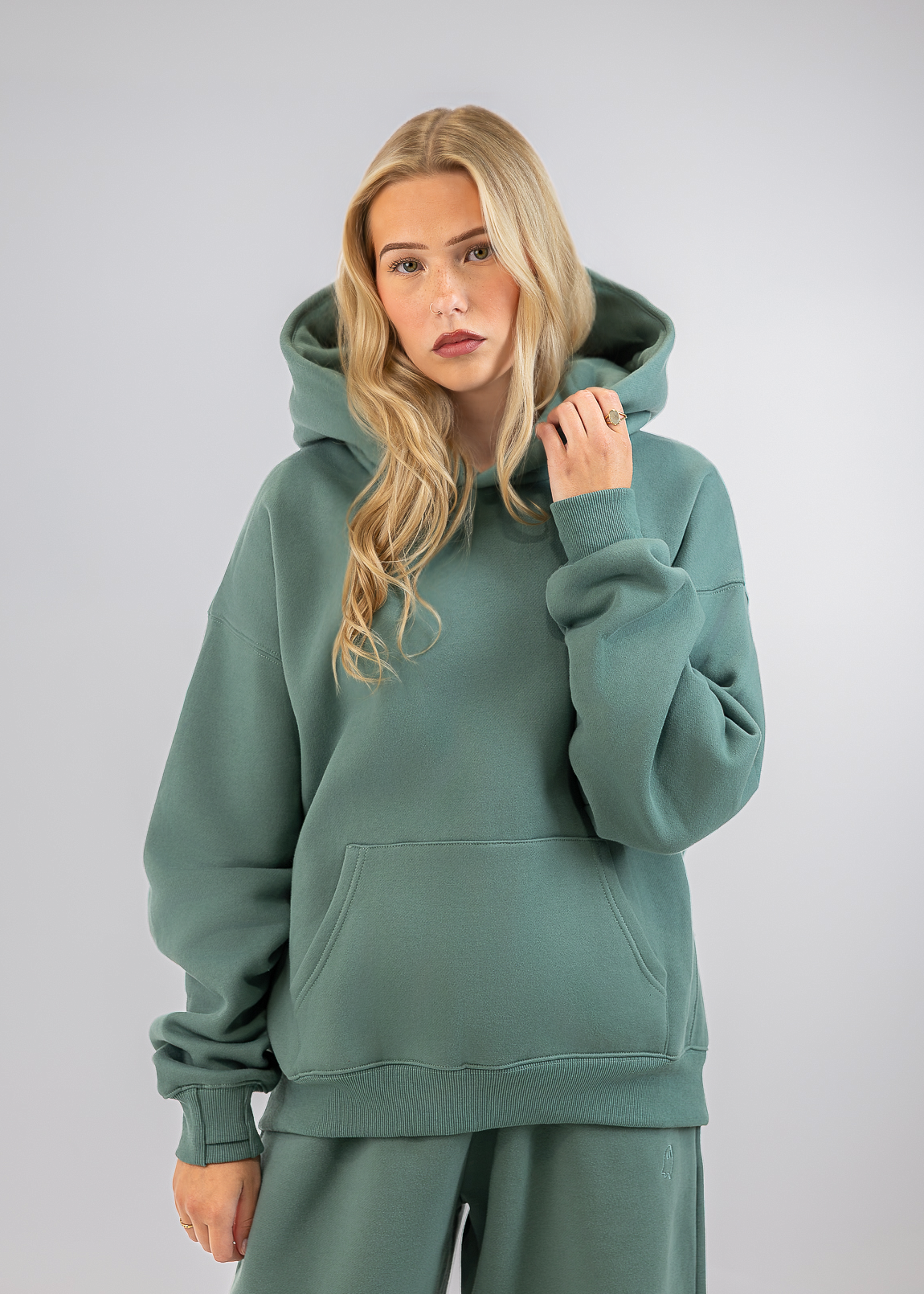 A blonde woman modeling a sage green hooded sweatshirt and matching sweatpants against a light gray studio background.