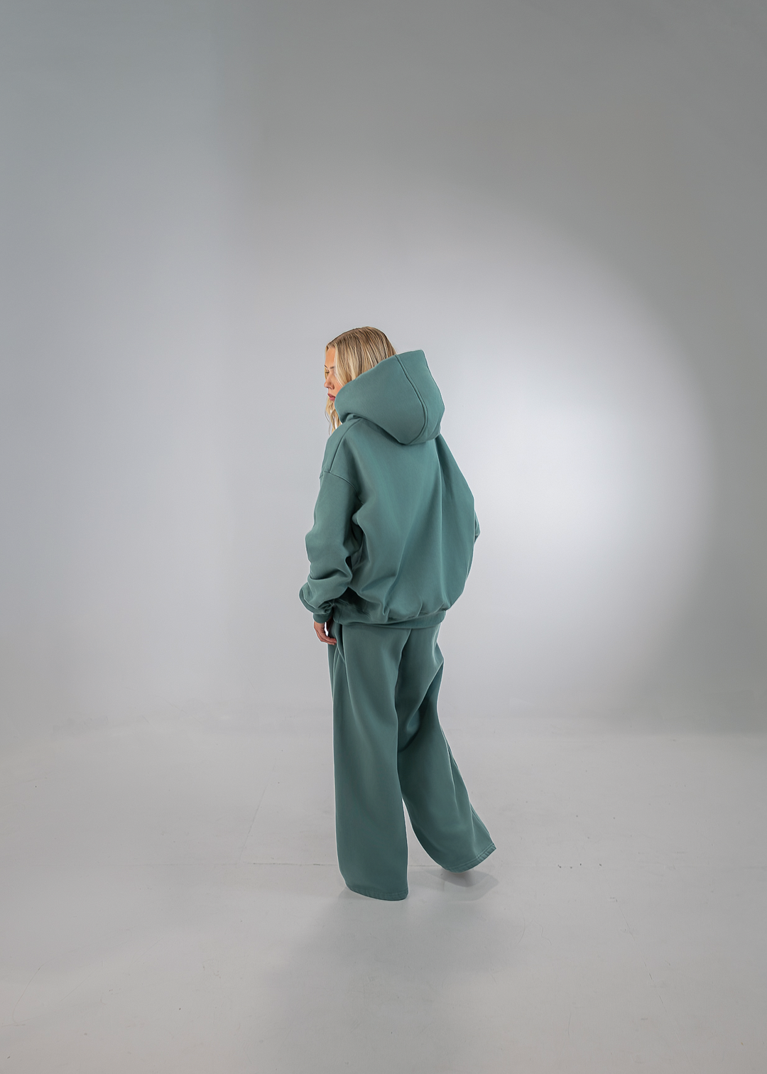 A child wearing a matching teal-green hooded sweatshirt and wide-leg sweatpants, standing in profile against a light gray studio background.