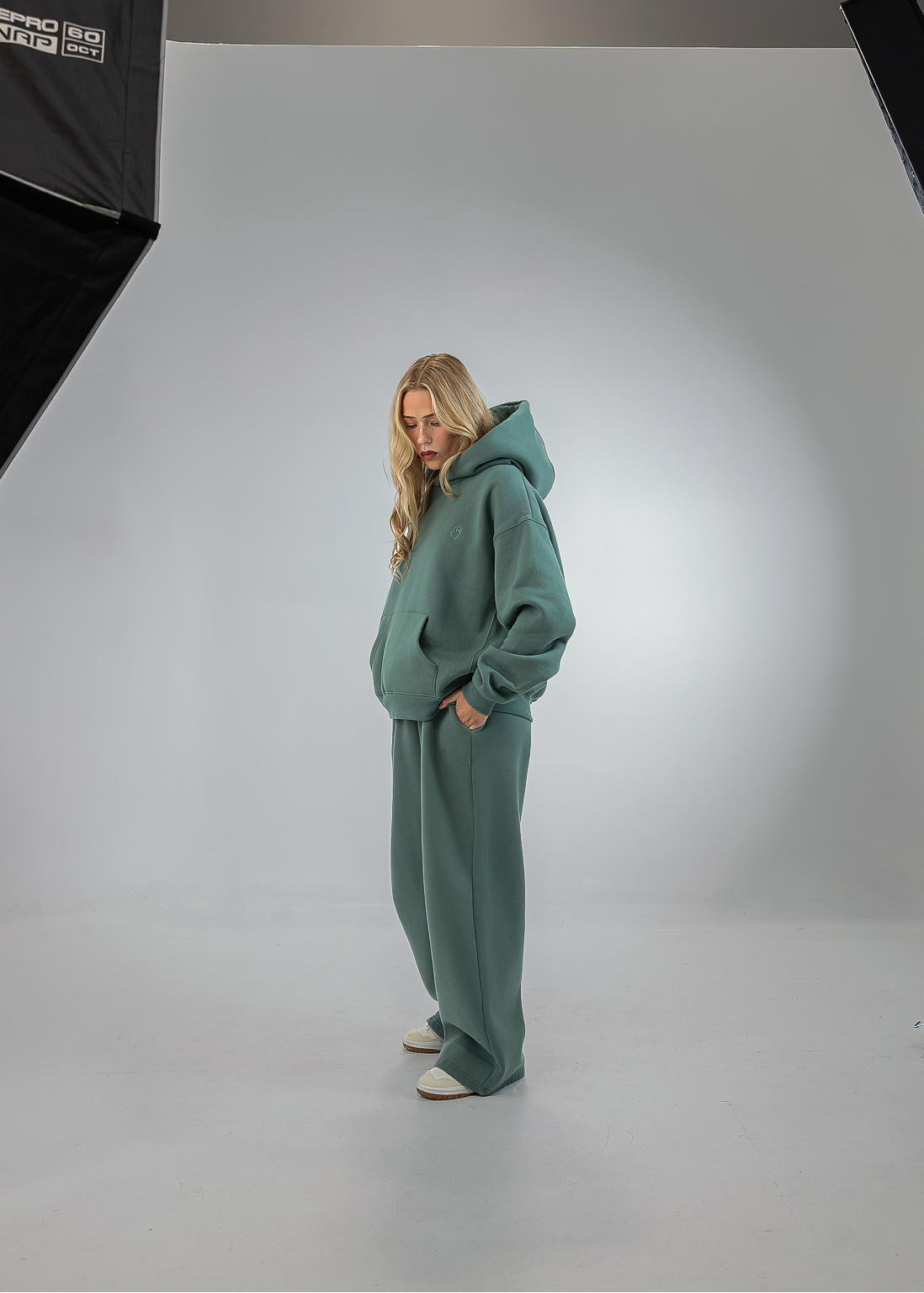 A woman stands in a white studio setting wearing a matching sage green lounge set consisting of a hooded sweatshirt and wide-leg sweatpants. Studio softbox lights are visible on the left and right sides of the background.