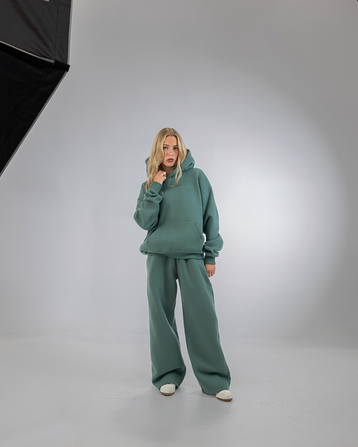 A woman modeling a matching sage green lounge set consisting of a pullover hoodie and wide-leg sweatpants, standing in a studio setting with softbox lights visible on the left and right.