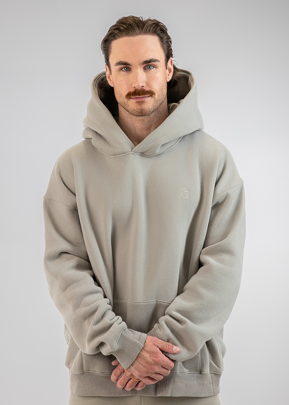 A man wearing a light gray oversized pullover hoodie with a matching pair of pants, standing against a neutral gray studio background.