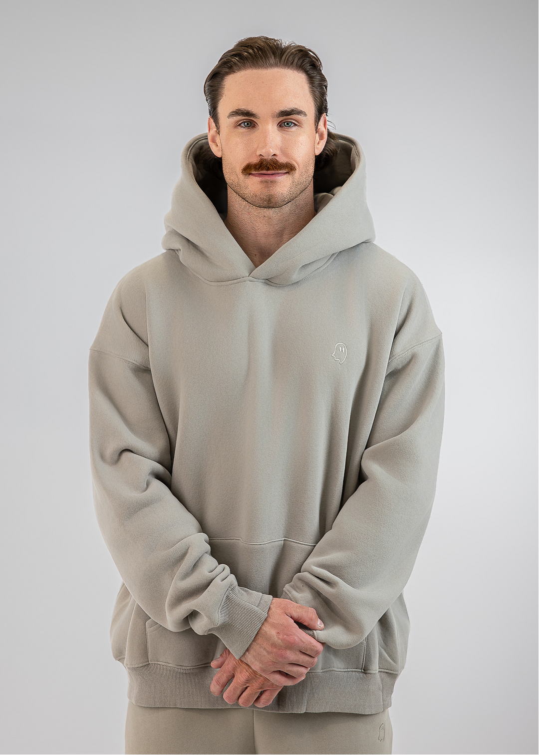 A man wearing a light gray oversized pullover hoodie with a small embroidered logo on the left chest, paired with matching light gray pants, standing against a neutral gray studio background.