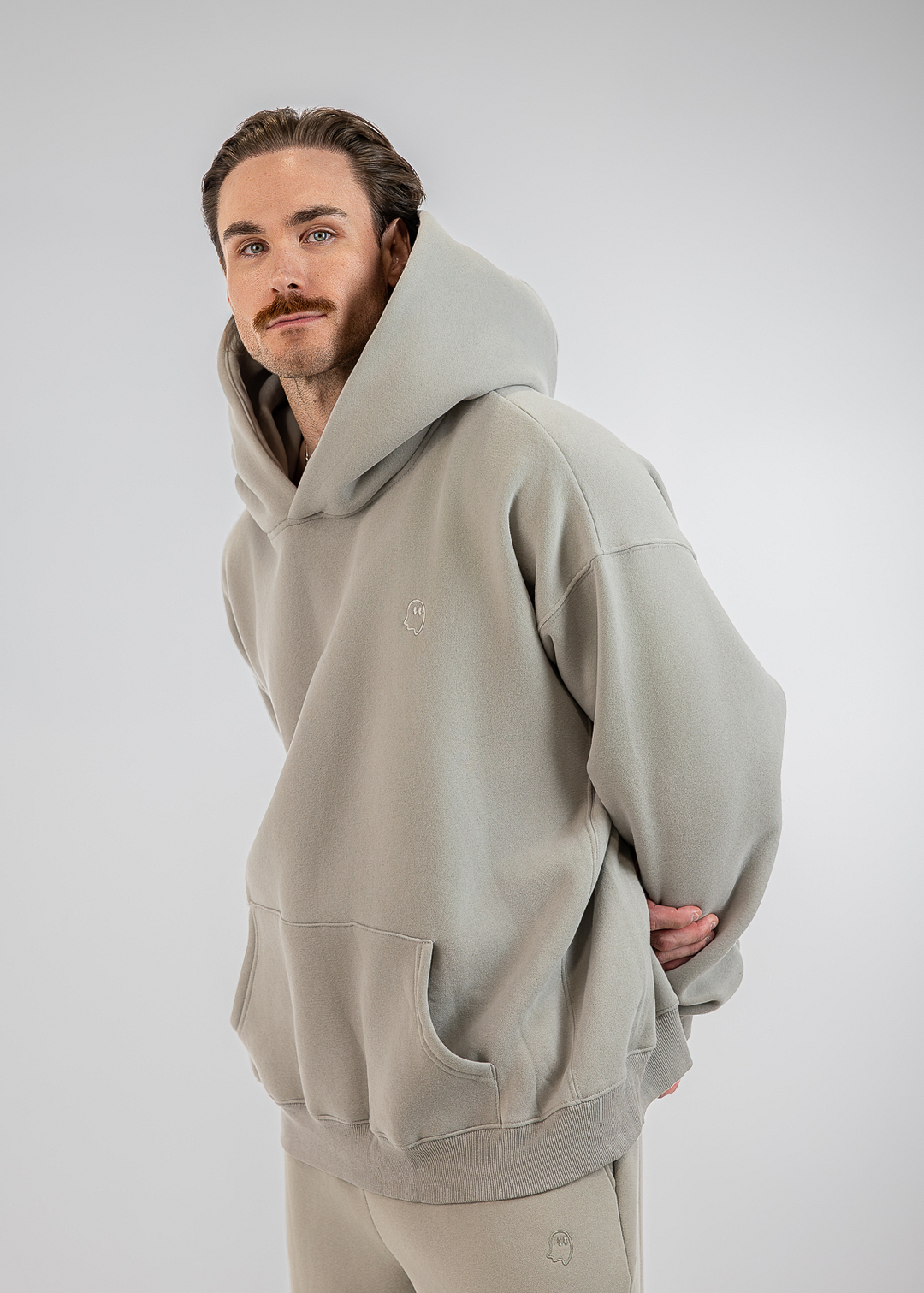 A man wearing a light gray oversized hoodie and matching sweatpants, photographed against a neutral light gray studio background.