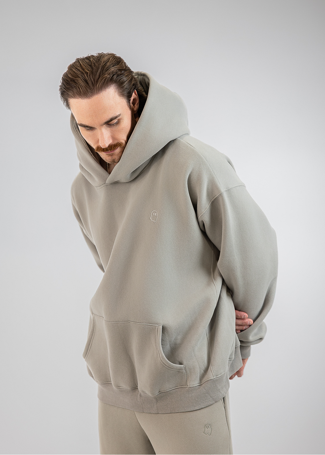 A man wearing a light gray oversized pullover hoodie and matching sweatpants, photographed against a neutral light gray studio background.
