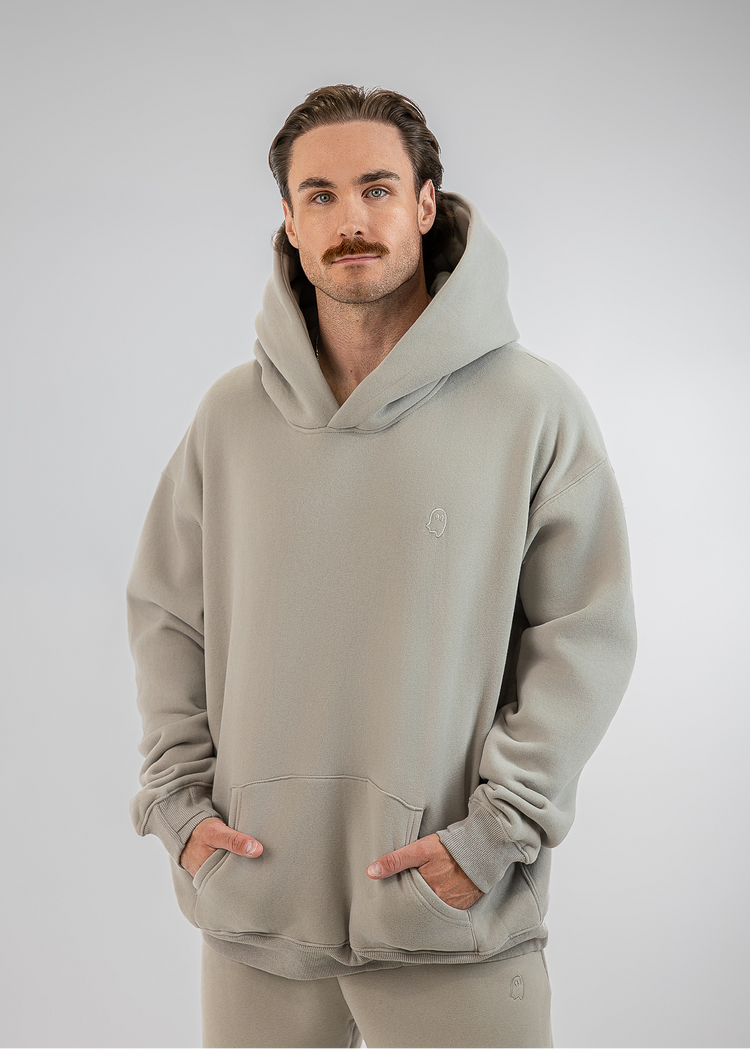 A man wearing a light gray hooded sweatshirt and matching sweatpants, standing against a neutral gray studio background.