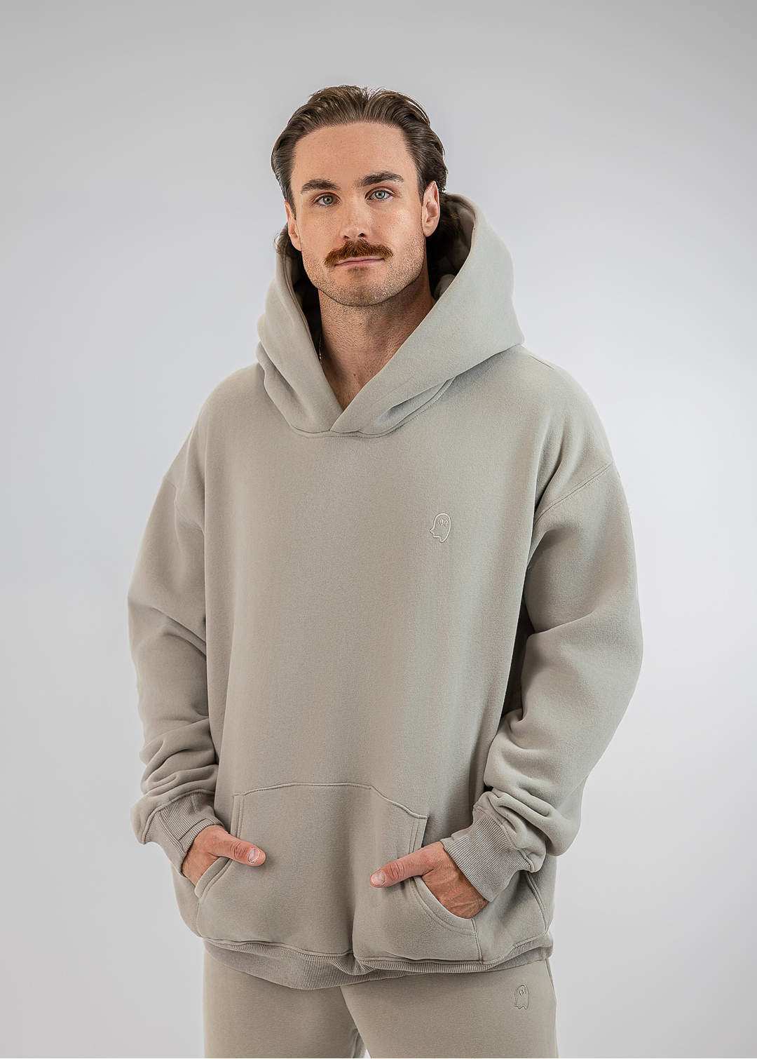 A man wearing a light gray hooded sweatshirt and matching sweatpants, standing against a neutral gray studio background.