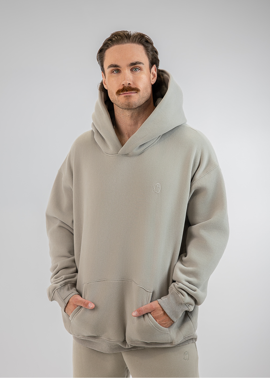 A man wearing a light gray hooded sweatshirt and matching sweatpants, standing against a neutral gray studio background.