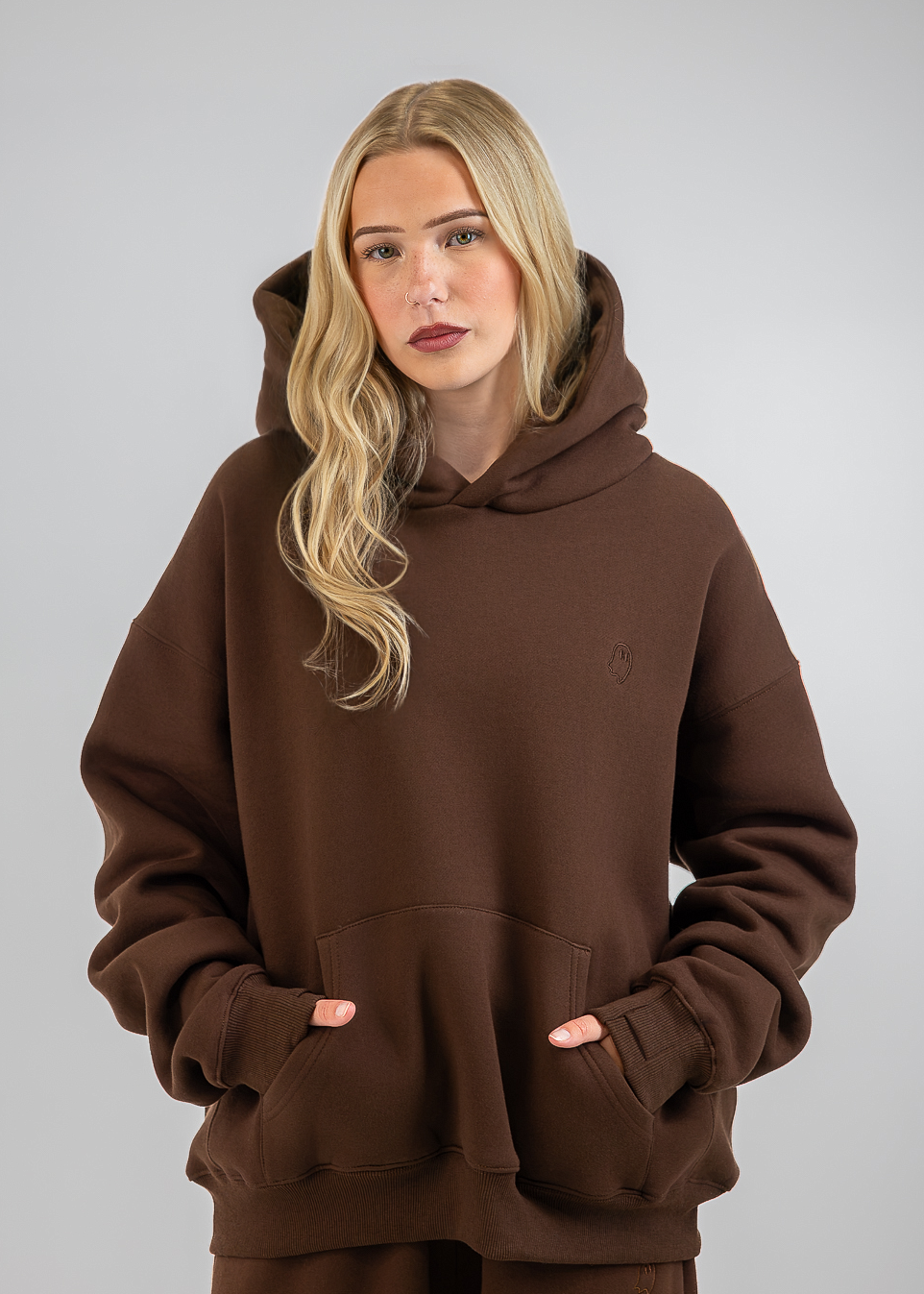 A blonde woman wearing an oversized brown hoodie and matching brown sweatpants, standing against a neutral gray studio background.