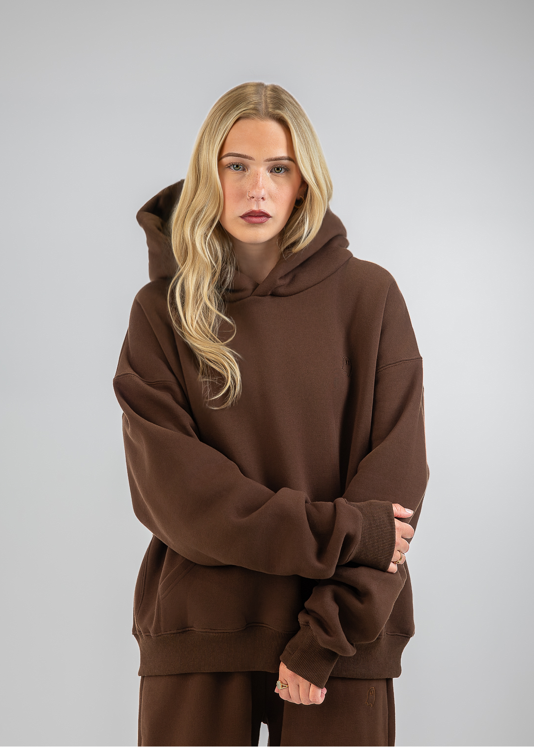 A blonde woman modeling an oversized brown hoodie and matching brown sweatpants against a neutral gray studio background.