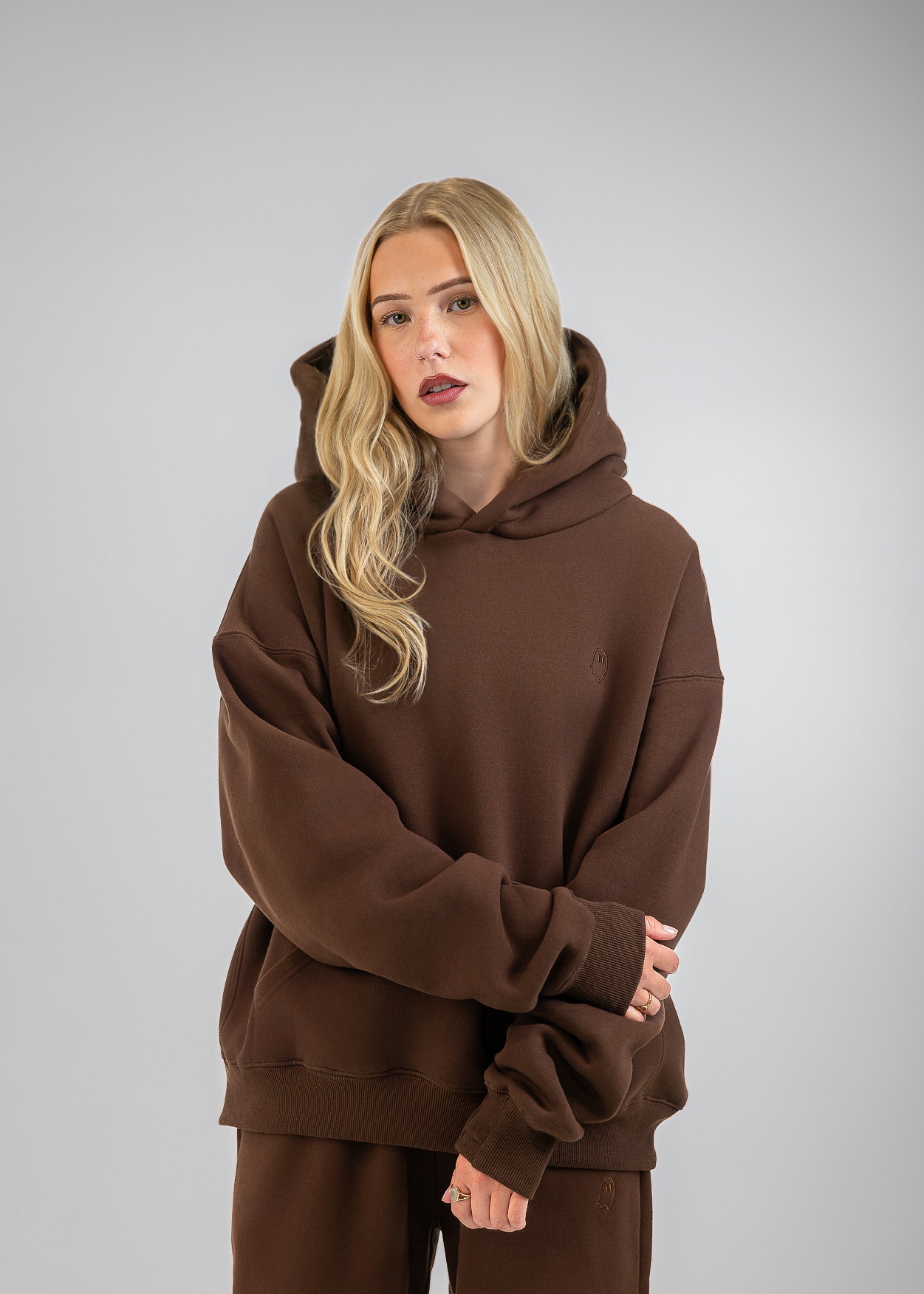 A blonde woman wearing an oversized brown hoodie and matching brown sweatpants, standing against a neutral gray studio background.