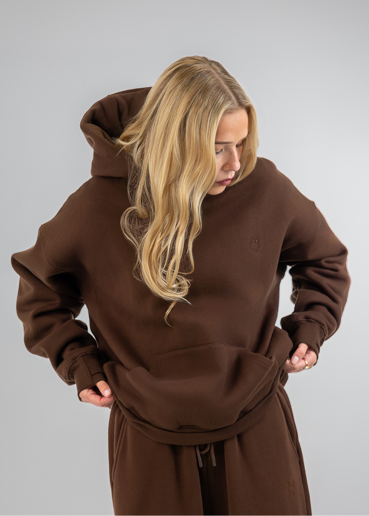 A woman wearing a matching brown hoodie and sweatpants set, looking down with her hands on the front of the hoodie. The background is a solid light gray studio backdrop.