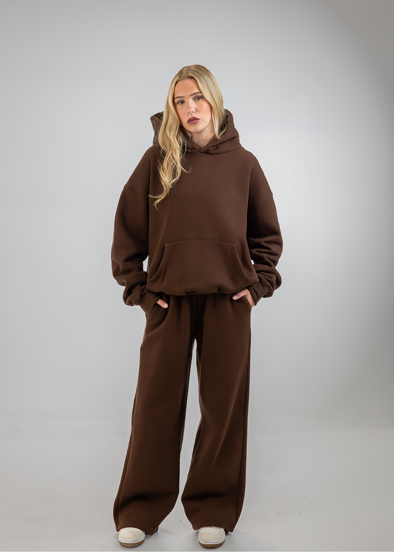 A woman modeling a matching brown lounge set consisting of a pullover hoodie and wide-leg sweatpants, standing against a neutral gray studio background.