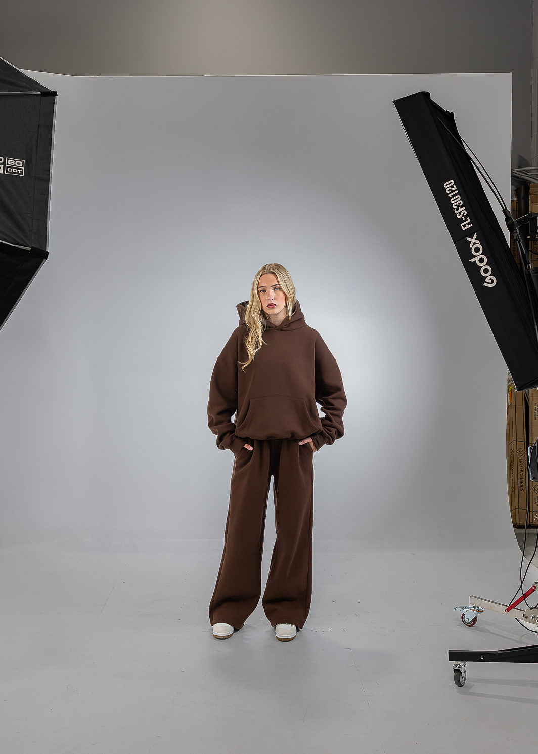 A woman stands in a studio wearing a brown sweatshirt and matching wide-leg pants, with a white seamless backdrop and softbox lights visible on both sides.