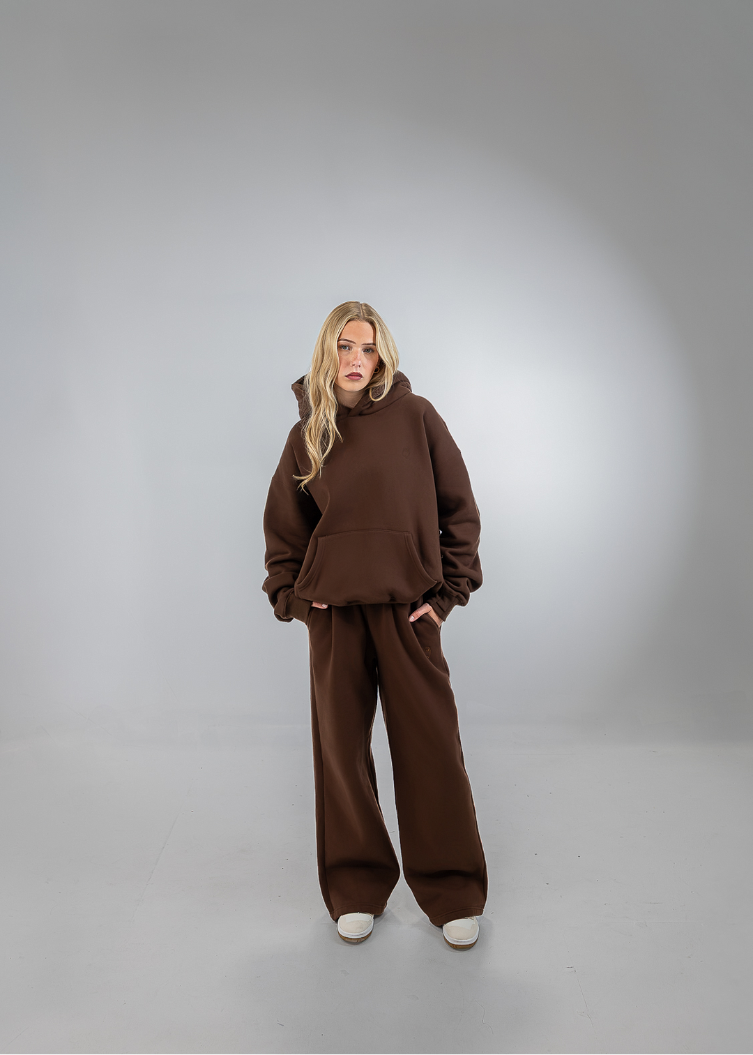 A woman stands in a studio wearing a matching brown lounge set consisting of a cropped hoodie and wide-leg pants, paired with white sneakers.