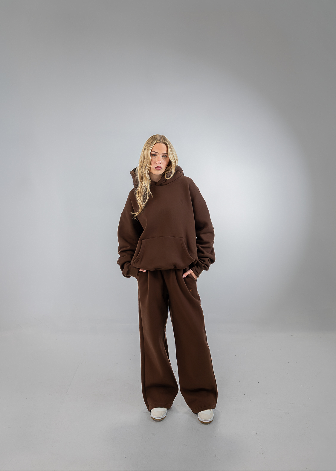 A woman stands in a studio wearing a matching brown lounge set consisting of a cropped hoodie and wide-leg pants, paired with white sneakers.