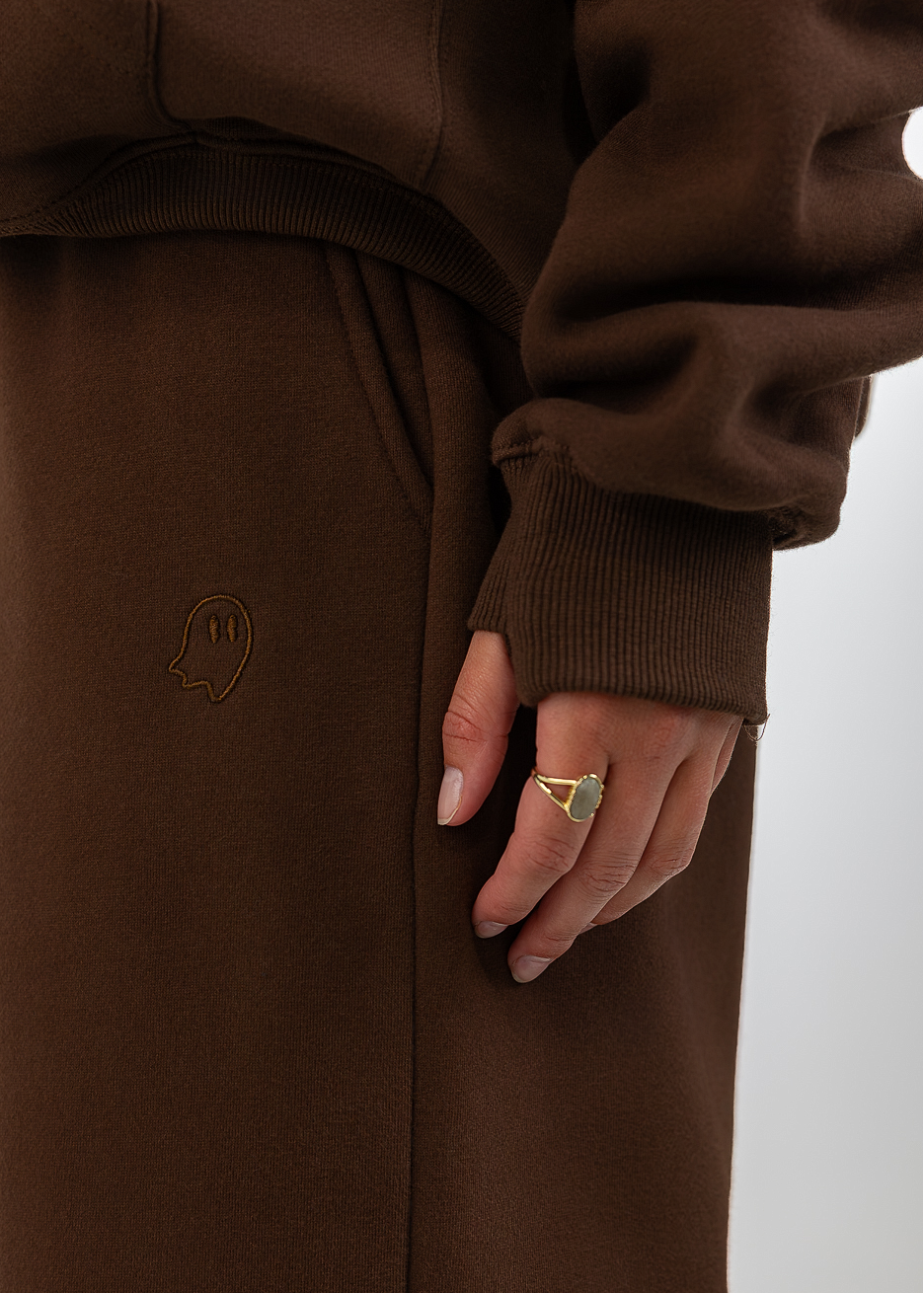 A close-up of a person wearing a matching brown sweatshirt and sweatpants set, with the hand resting on the thigh. The sweatpants feature a small embroidered logo on the left leg.