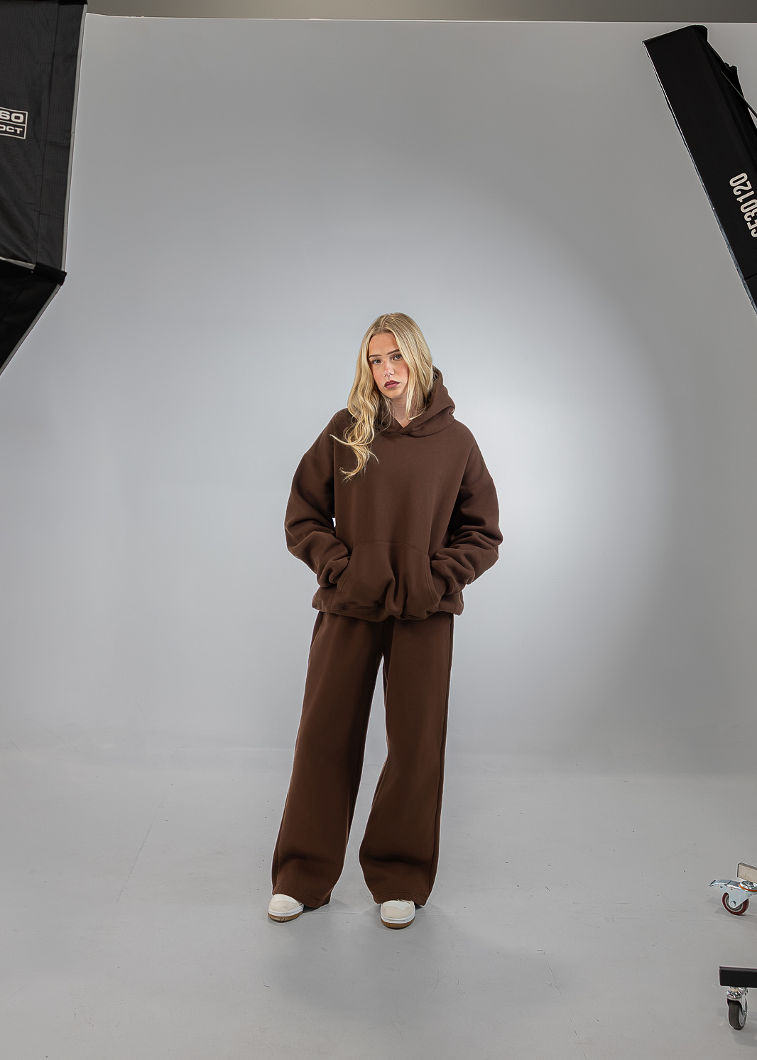 A woman stands in a studio wearing a matching brown hoodie and wide-leg sweatpants set, with studio lights visible on both sides of the white seamless backdrop.