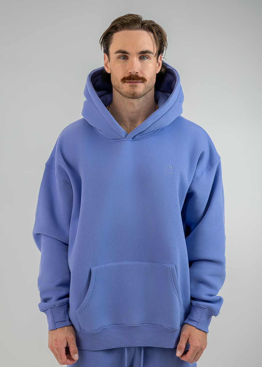 A man wearing an oversized light blue pullover hoodie with a kangaroo pocket and matching sweatpants, standing against a neutral gray studio background.