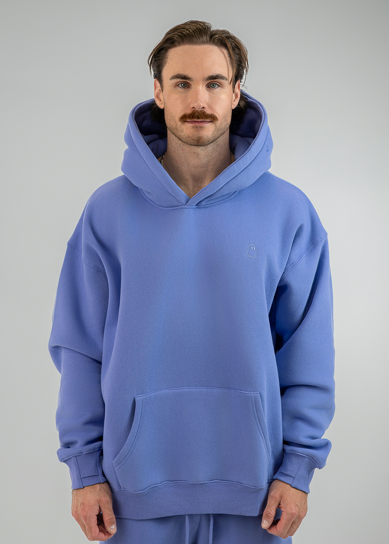 A man wearing an oversized light blue pullover hoodie with a kangaroo pocket and matching sweatpants, standing against a neutral gray studio background.