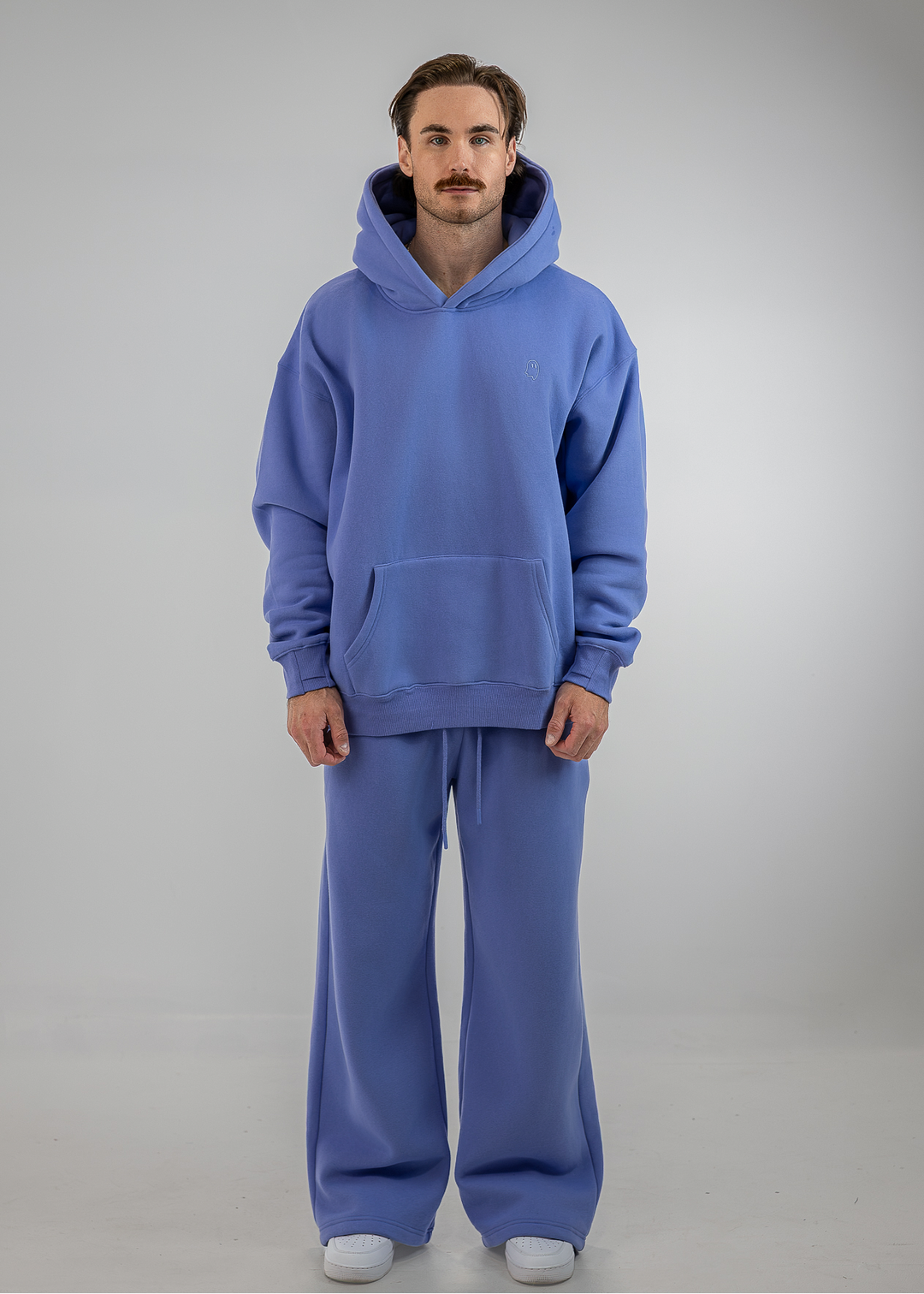 A man wearing a matching blue sweatshirt and wide-leg sweatpants set, standing against a neutral gray studio background.