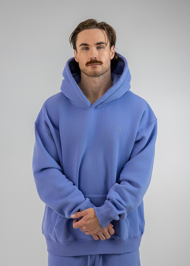 A man wearing a matching blue hoodie and sweatpants set, standing against a neutral gray studio background.