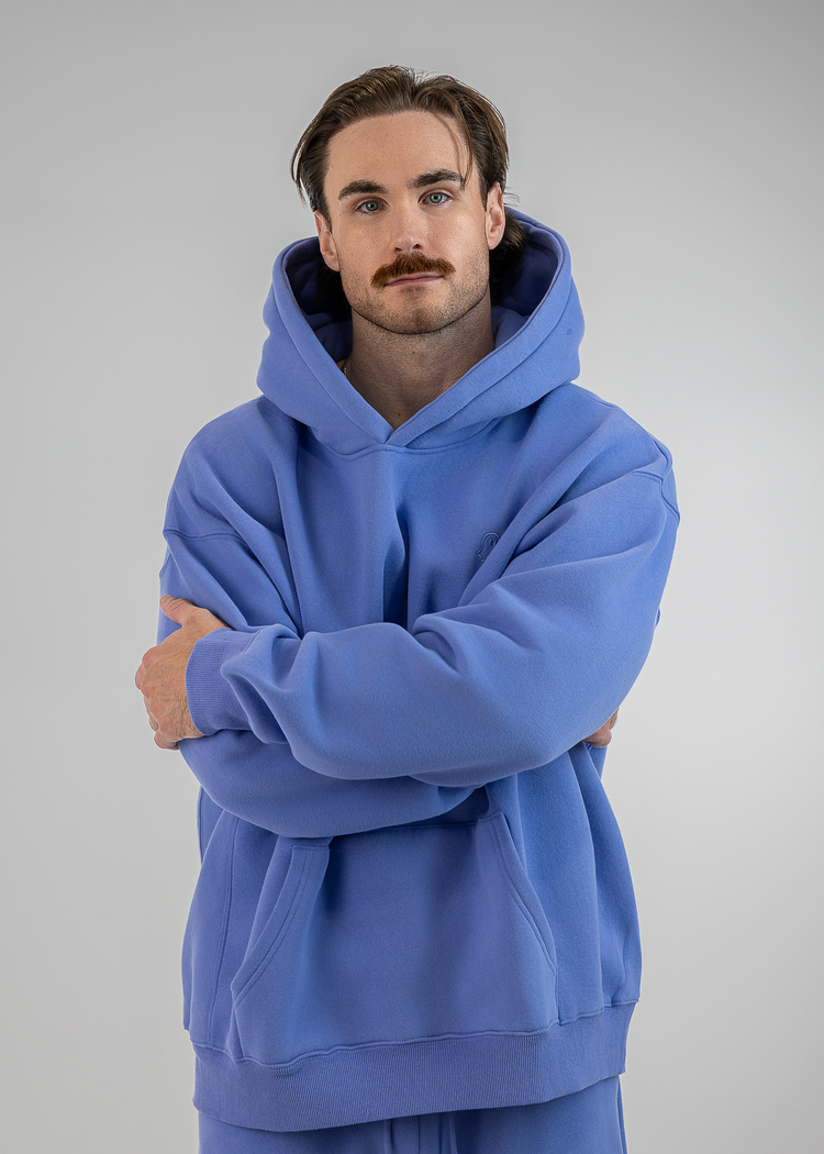 A man wearing an oversized blue hoodie and matching sweatpants, standing with arms crossed against a plain light gray studio background.