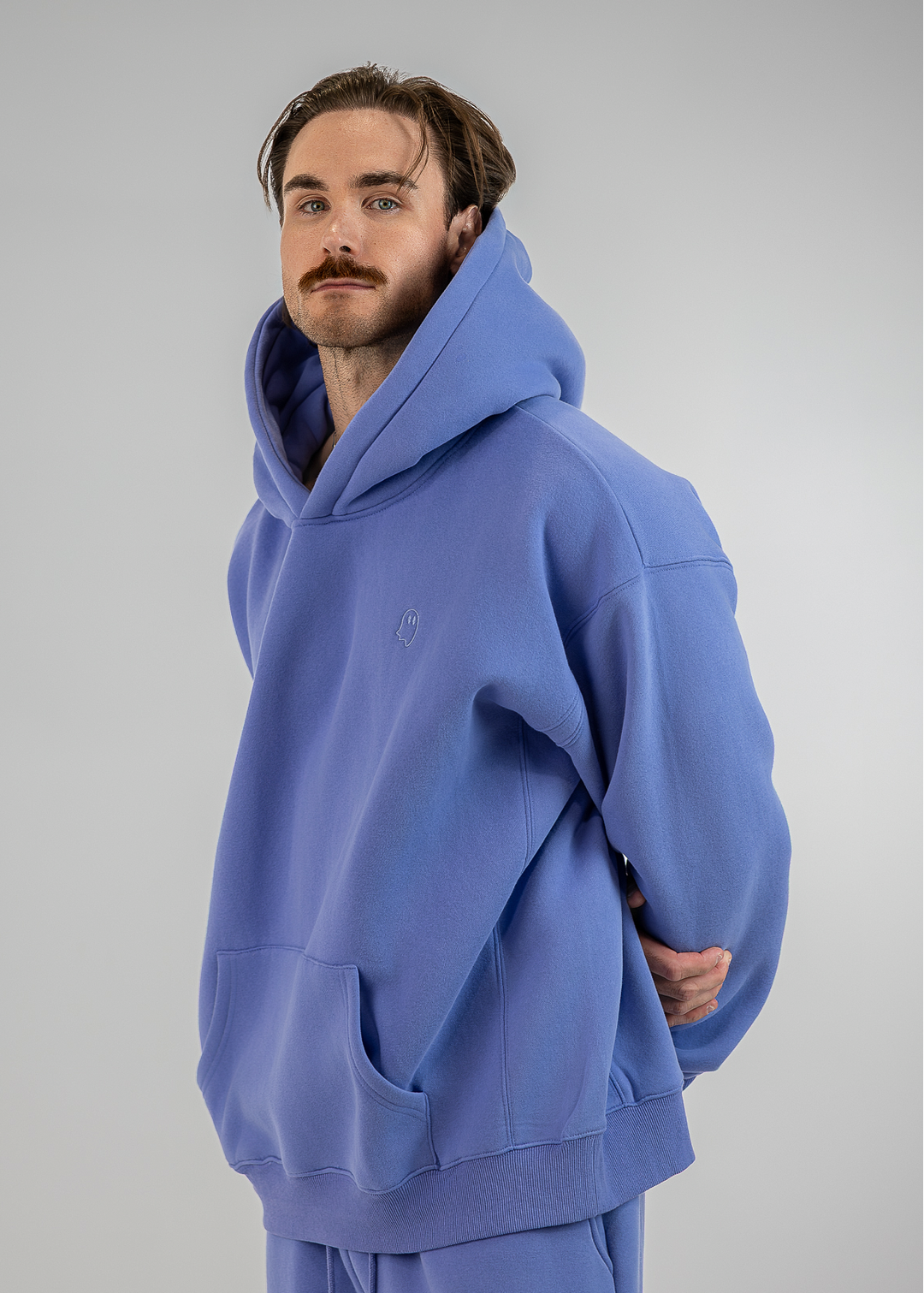 A man modeling a matching lavender-blue hoodie and sweatpants set against a plain light gray studio background.
