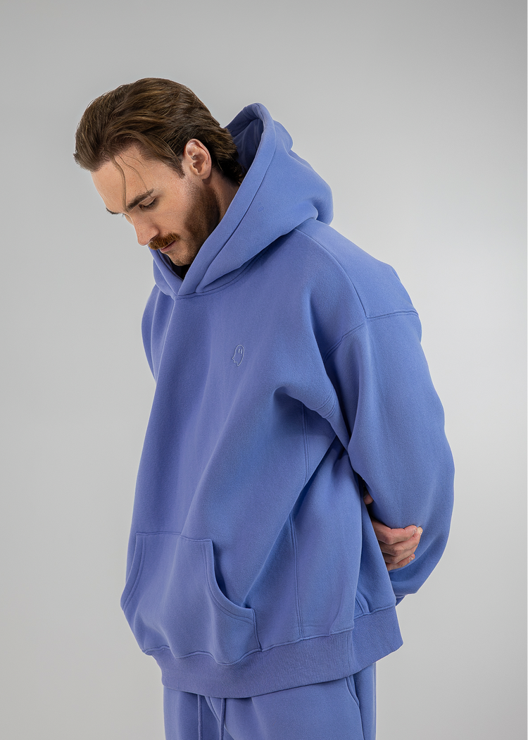 A man wearing an oversized blue hoodie and matching sweatpants, photographed against a plain light gray studio background.