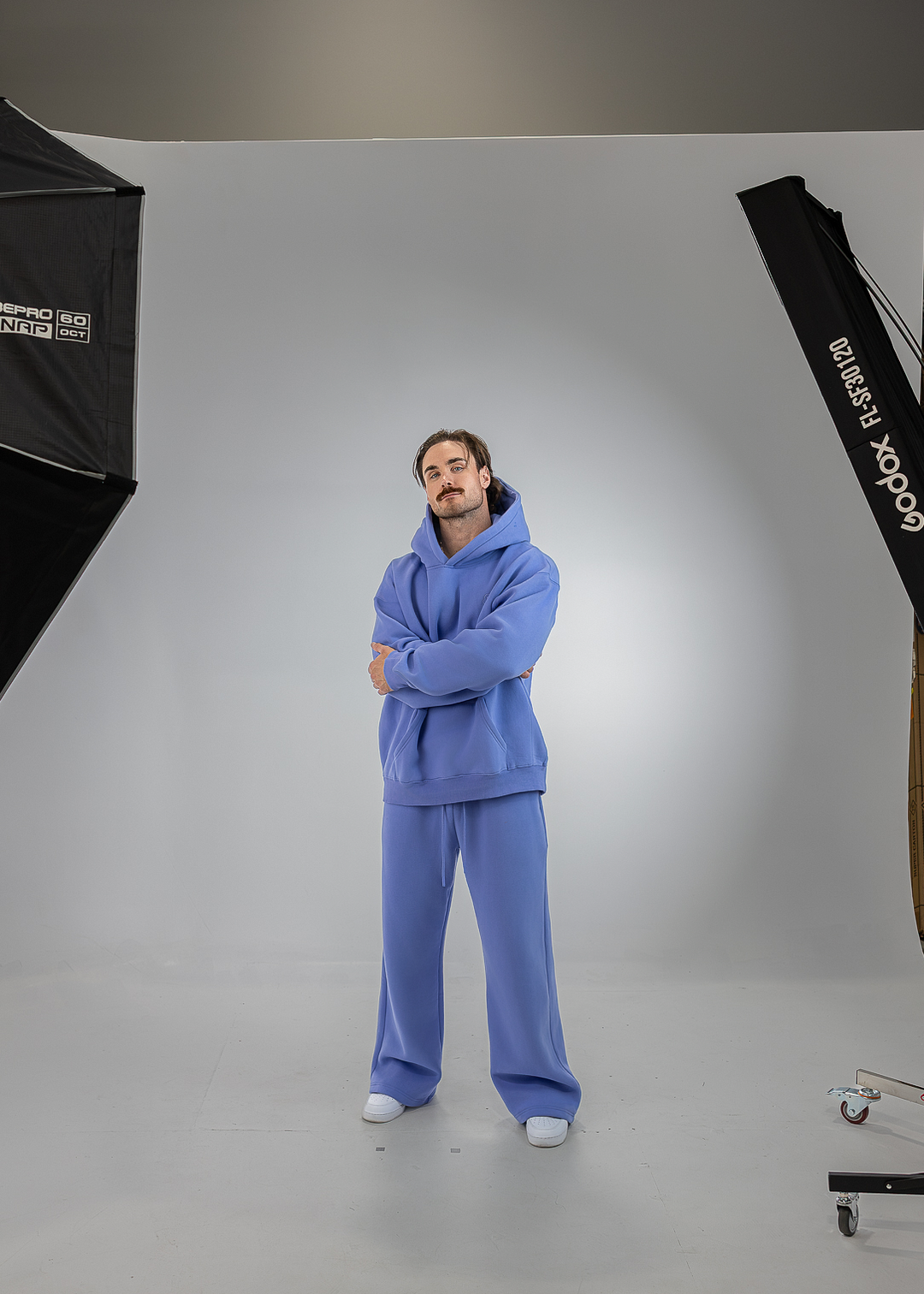 A man stands in a studio wearing a matching lavender-purple sweatshirt and sweatpants set. The studio setup includes a white seamless backdrop, a softbox light on the left, and another light stand on the right.