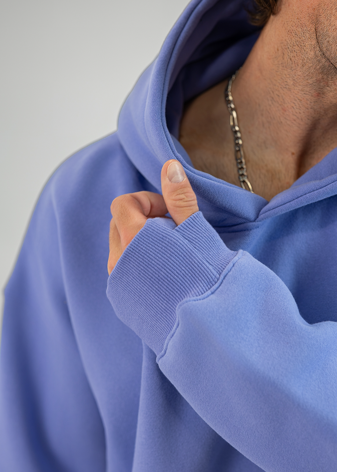Close-up of a person wearing a periwinkle blue hoodie, pulling the hood down slightly with one hand. The person is also wearing a silver chain necklace.