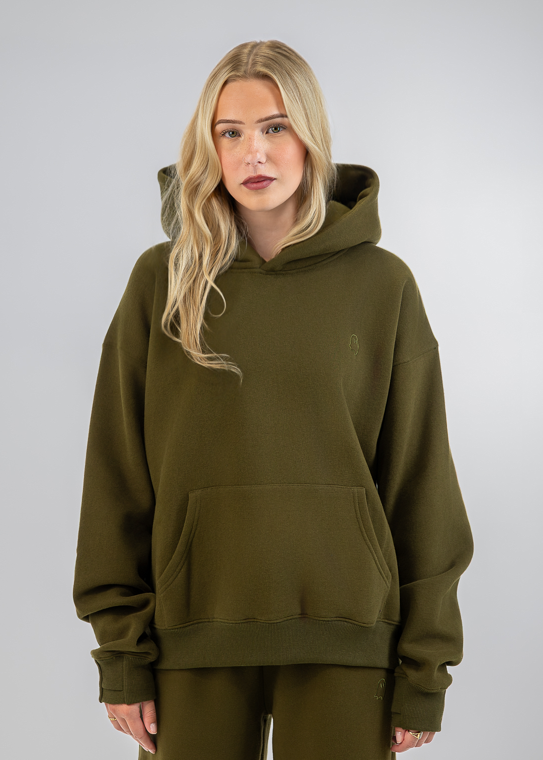 A blonde woman wearing an oversized olive green pullover hoodie with a kangaroo pocket and matching olive green sweatpants, standing against a neutral light gray studio background.