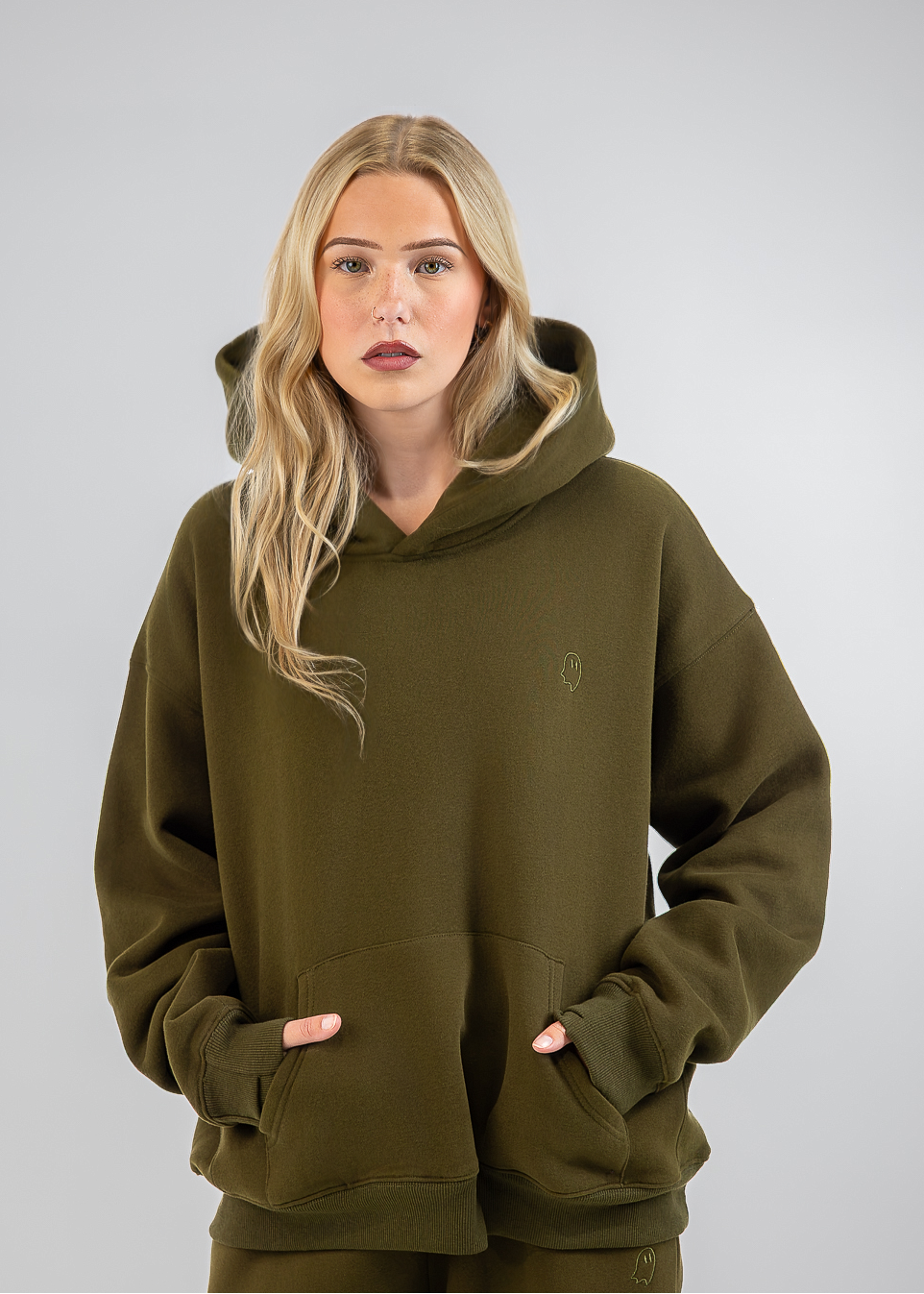 A blonde woman modeling an olive green pullover hoodie and matching sweatpants against a neutral gray studio background.