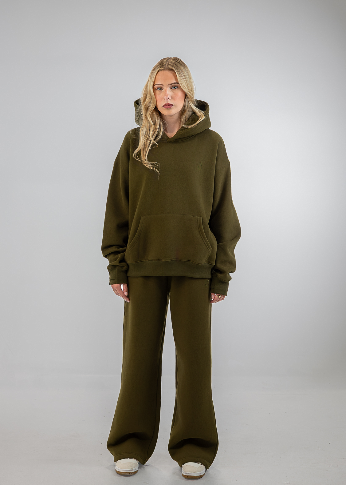 A blonde model stands against a light gray studio background wearing an oversized olive green hoodie and matching wide-leg sweatpants.