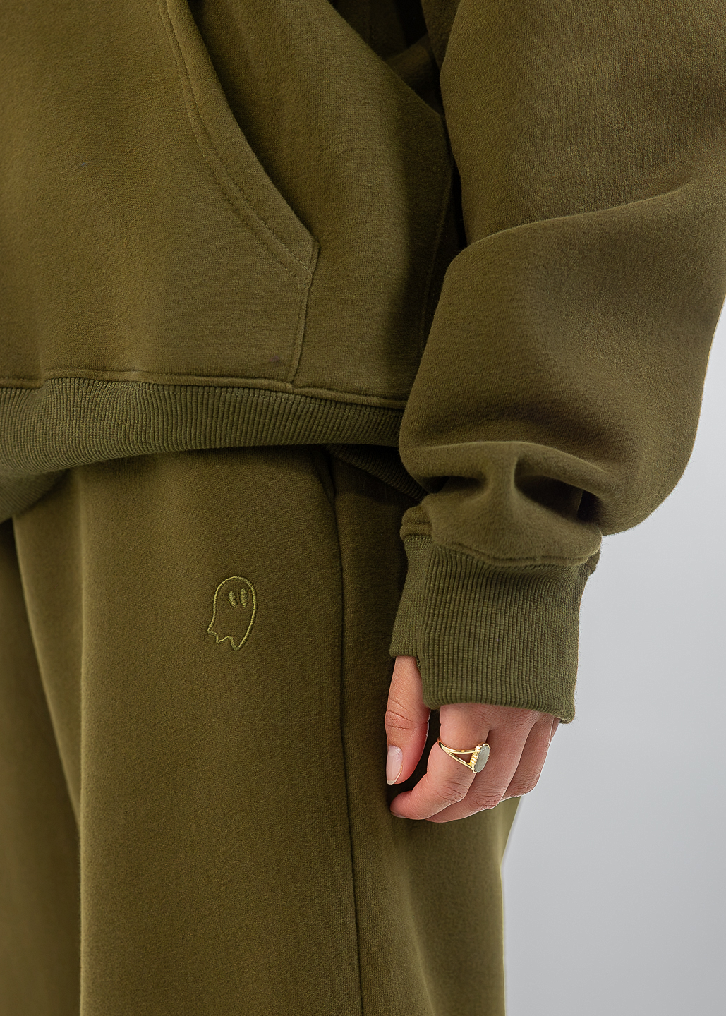 Close-up of a person wearing an olive green sweatshirt and matching sweatpants, with a small embroidered ghost logo on the thigh and a gold ring on one finger, against a light gray background.