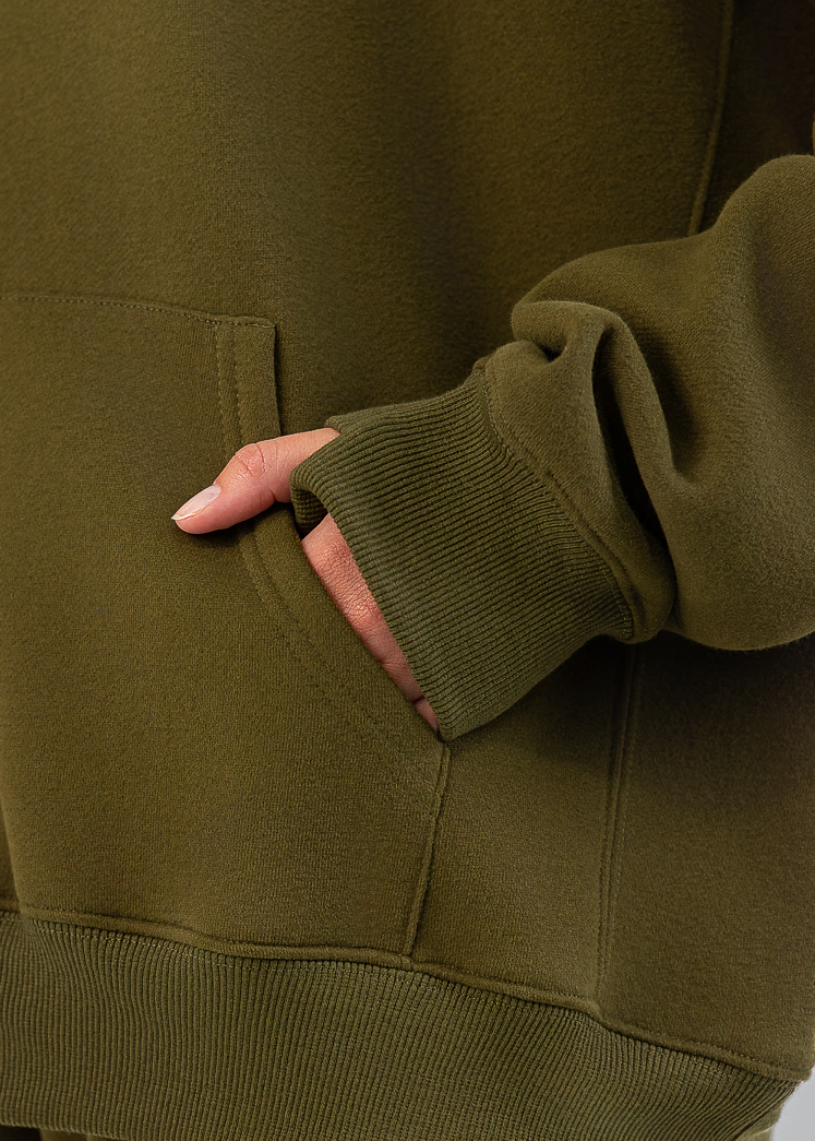 Close-up of a person wearing an olive green sweatshirt and matching sweatpants, with one hand in the sweatshirt pocket and a small embroidered logo on the thigh.