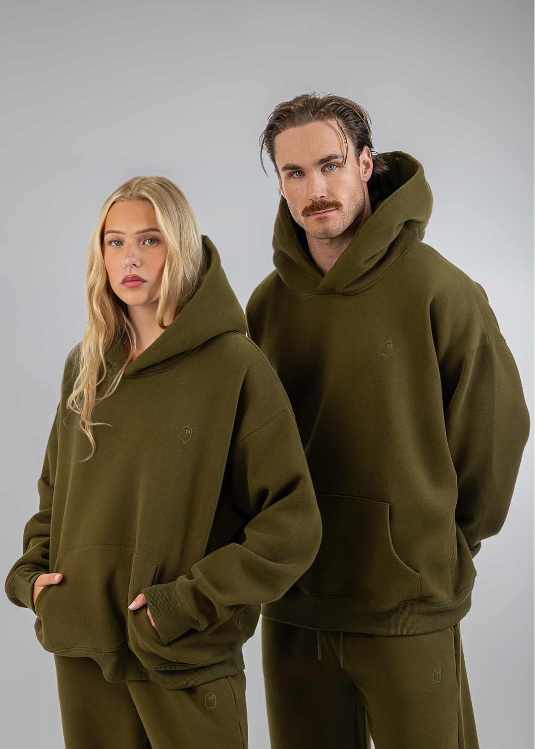 A man and a woman modeling matching olive green hoodies and sweatpants sets against a neutral gray studio background.