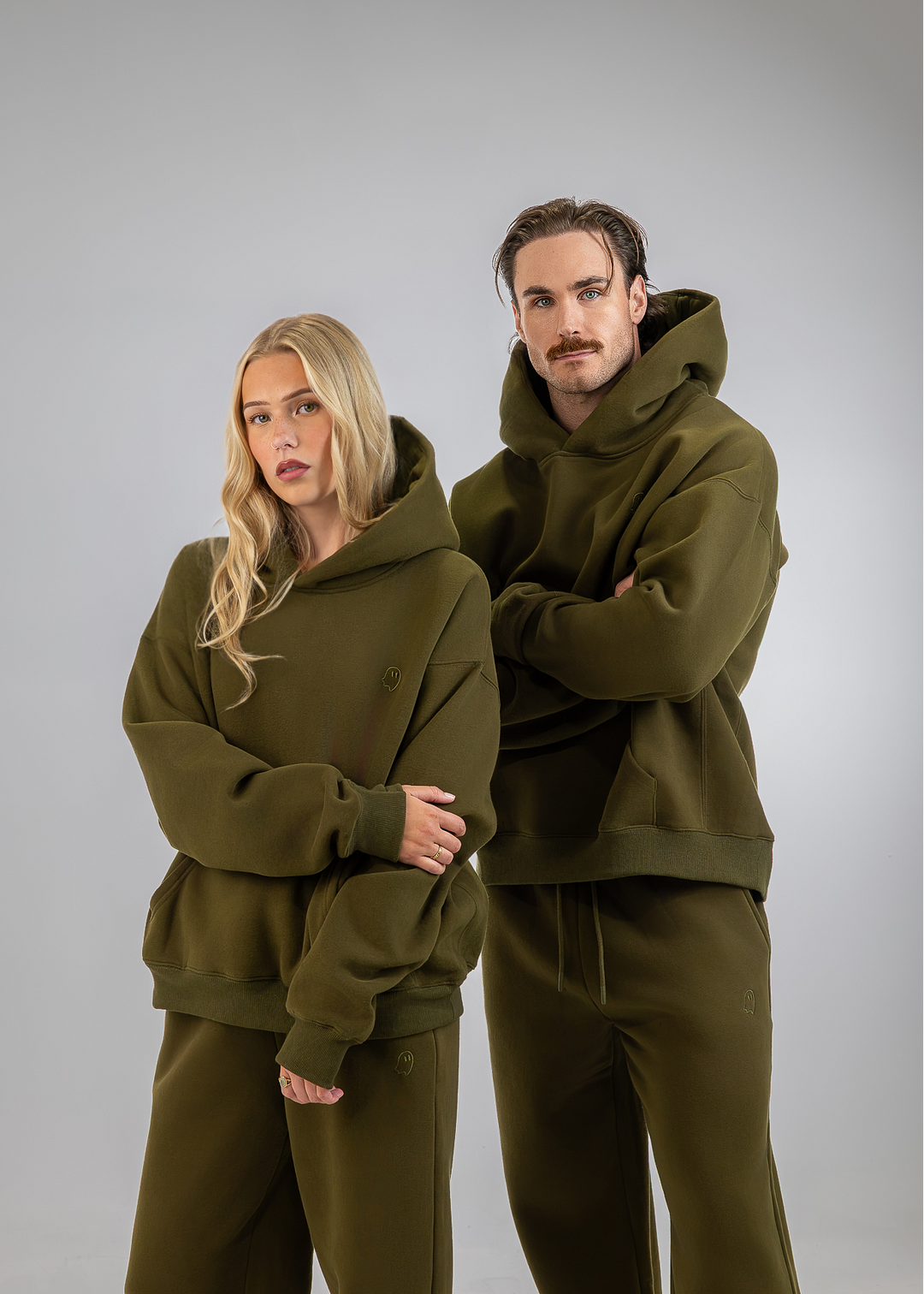 A man and a woman modeling matching olive green hoodies and sweatpants sets against a solid gray studio background.
